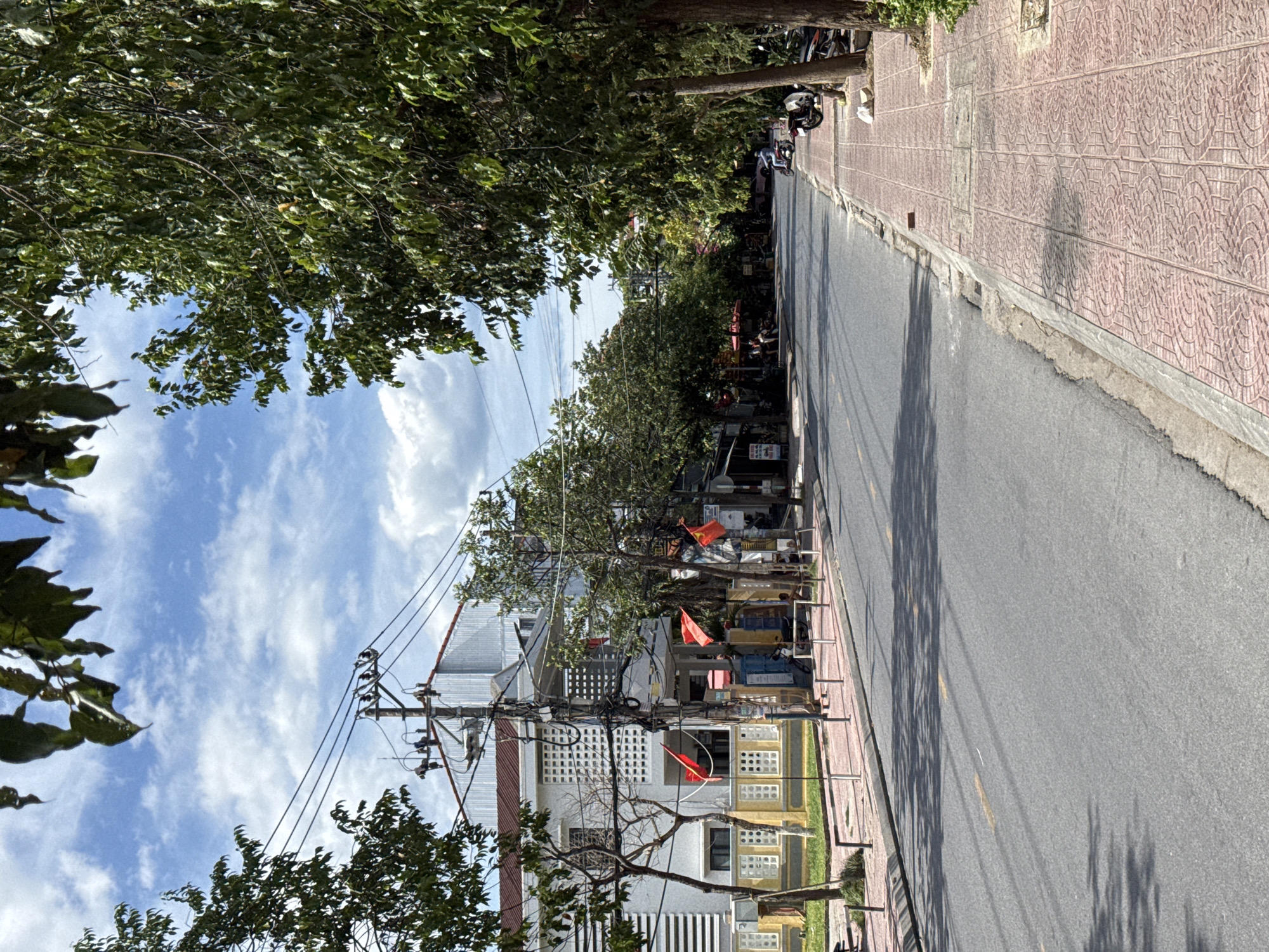 This is an eye-level shot of a quiet street in Hoi An, Vietnam. The street runs diagonally from the bottom right to the top left of the frame, bordered by a sidewalk on the right and a building on the left. The road is paved with asphalt and has a yellow line marking the center. The sidewalk is paved with reddish-pink tiles arranged in a decorative pattern.
On the left side of the street, a white building with a red-tiled roof is visible. It has a light-colored facade with white-grated windows and yellow decorative elements near the ground. Red Vietnamese flags are displayed on poles in front of the building. A utility pole with numerous wires runs along the sidewalk in front of the building. Trees with green foliage line the street on both sides, casting shadows on the road and sidewalk.
On the right side of the street, more trees are visible, and parked motorcycles are present. A person is seen riding a motorcycle down the street in the distance. The sky is blue with scattered white clouds. The scene appears to be captured during daylight, under a bright sky, suggesting a warm, possibly humid, day. The overall atmosphere is calm and tranquil, with minimal activity.