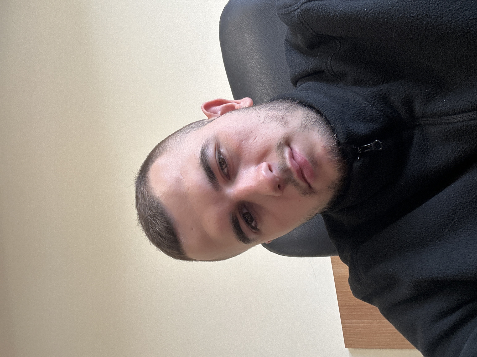 A close-up, selfie-style, vertically-oriented photograph shows a fair-skinned male with a buzzcut. He has light brown eyes and a light beard. He is wearing a black fleece jacket. He appears to be seated in a black chair. The background consists of a light beige wall and a portion of a light brown wooden surface. The image does not contain information that would indicate the location is Plovdiv, Bulgaria.