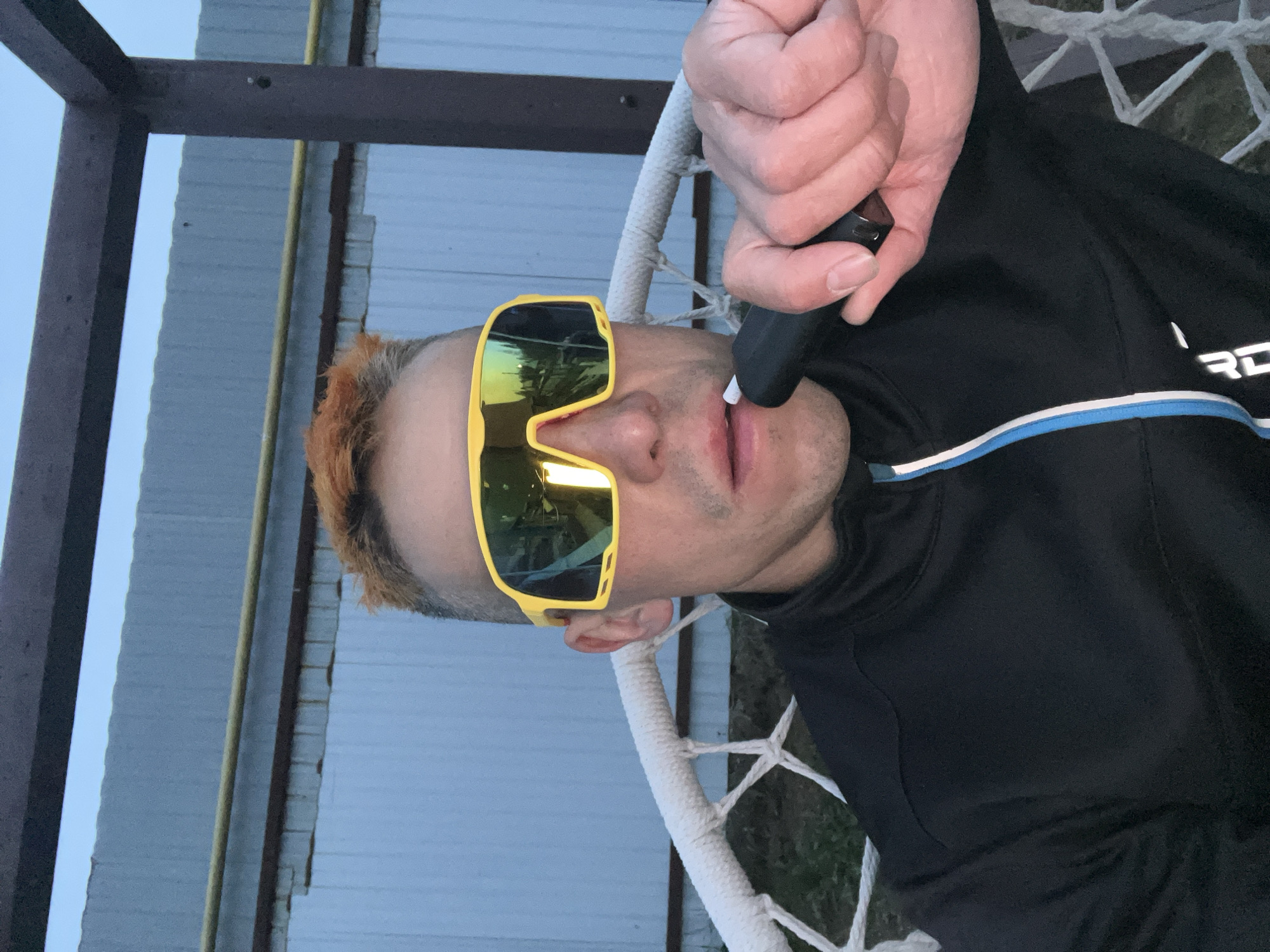 A man is seated in a white rope chair, wearing yellow sunglasses and a black jacket with a blue and white stripe. He is holding a black object to his mouth. The setting appears to be outdoors, possibly in Chaikovsky, Russia, with a metal structure and corrugated metal fencing visible in the background. The sky is overcast.