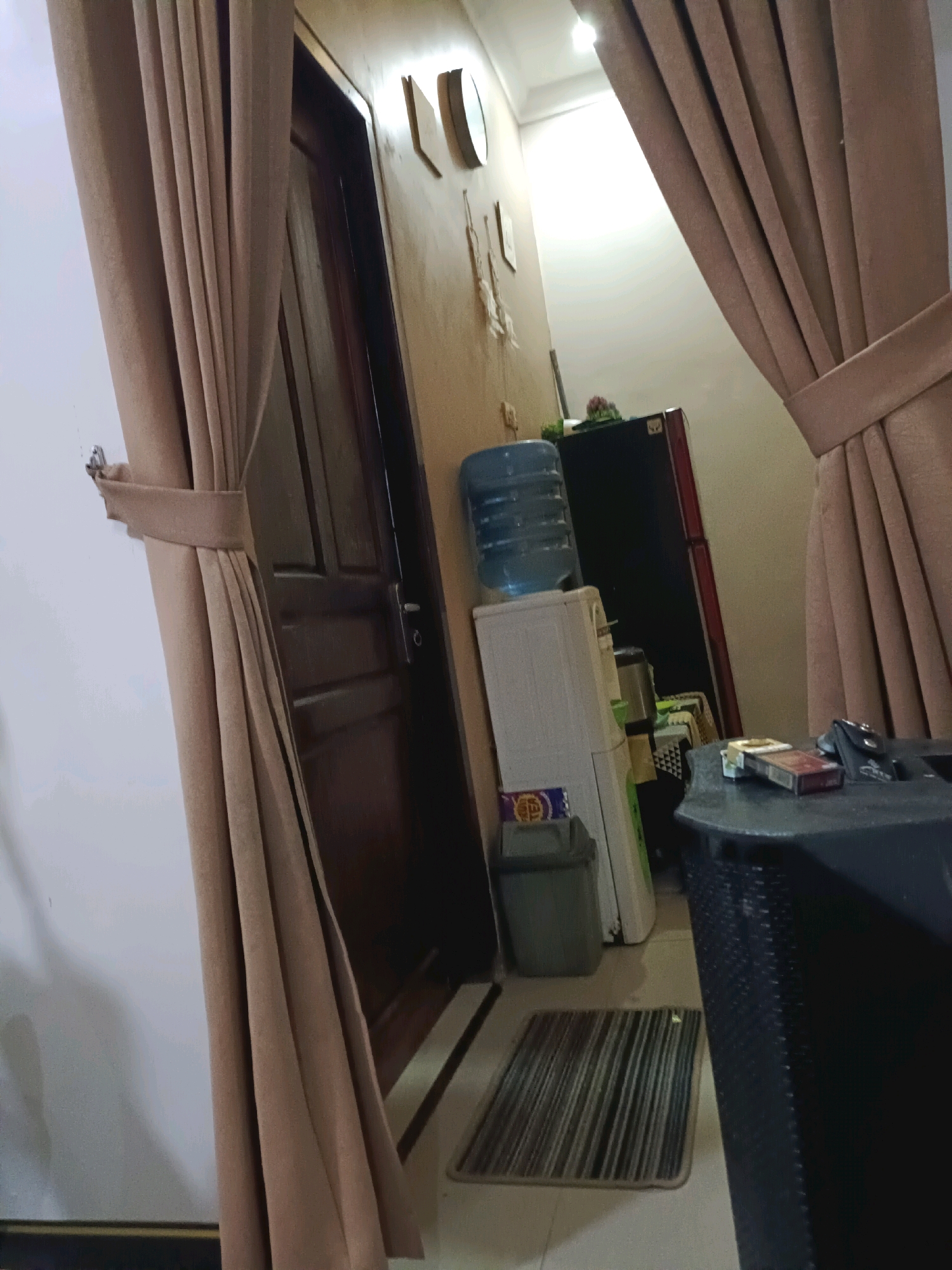 The image depicts an interior scene, possibly a hallway or common area within a residence in Rancang Kencono, Indonesia. The foreground is dominated by a large, tan-colored curtain tied back with a matching band, obscuring a portion of the view. To the left, a white wall is visible.
Further into the scene, a dark brown wooden door is partially open, revealing its paneling and a silver doorknob. Adjacent to the door, a beige wall displays two decorative items: a circular clock and a framed picture or plaque.
In the middle ground, a white water dispenser with a large blue water bottle on top stands to the left. Next to it is a dark gray trash can. Behind the water dispenser, a black refrigerator with a red stripe is visible.
To the right of the refrigerator, a dark gray table with a textured surface is present. On the table, a pack of cigarettes and a small black pouch are placed. A striped mat lies on the tiled floor in front of the water dispenser and trash can. The overall lighting suggests it is daytime, with a bright light source coming from above and to the right.