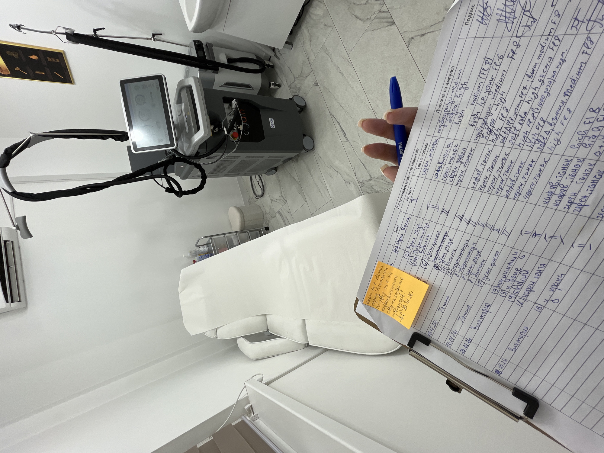 The image captures a clinical setting, likely a dermatology or cosmetic treatment room. Prominently visible is a modern laser hair removal machine, positioned near a white treatment bed. A person is holding a clipboard with a handwritten document and a pen in their hand, presumably patient records or treatment notes. A sticky note is attached to the side of the clipboard. The environment appears sterile, with a white color scheme dominating the walls and furniture. The presence of the laser machine, treatment bed, and patient records indicates a medical or cosmetic procedure is taking place or is about to. The location is unknown, but the language on the documents appears to be Bulgarian.