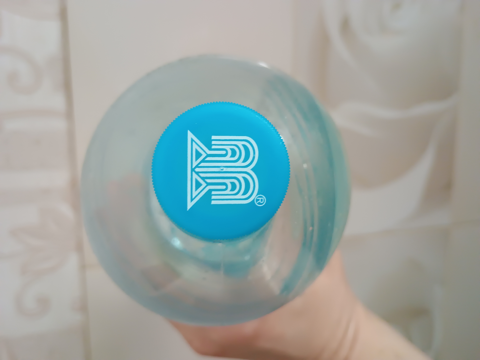 A close-up, overhead shot shows a hand holding a clear plastic bottle of water, its blue cap prominently displayed with a white logo. The background is a blurry, light-colored surface with a tiled pattern. The logo appears to be a stylized "W" with a triangle above it, and a registered trademark symbol to the lower left. The water inside the bottle is a light blue color.