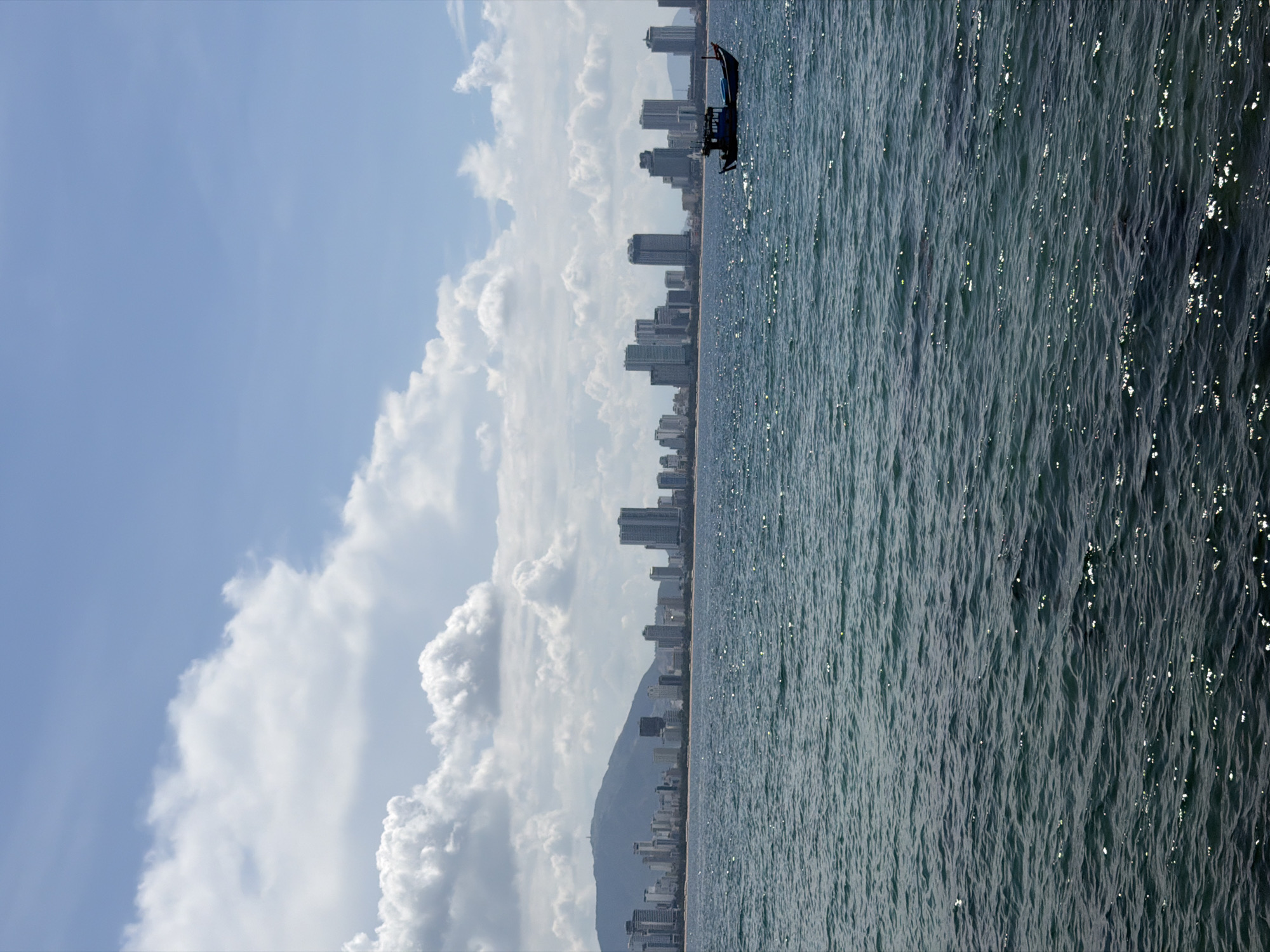 The image shows a coastal cityscape in Nha Trang Ward, Vietnam. A body of water, possibly the South China Sea, fills the right half of the frame, with sunlight reflecting off its surface. To the left is a view of the coastline, displaying a series of high-rise buildings along the beach. In the distance, a mountain or large hill can be seen. The sky is visible above, with a mix of clear blue and white, puffy clouds. A small boat is docked at the shoreline.