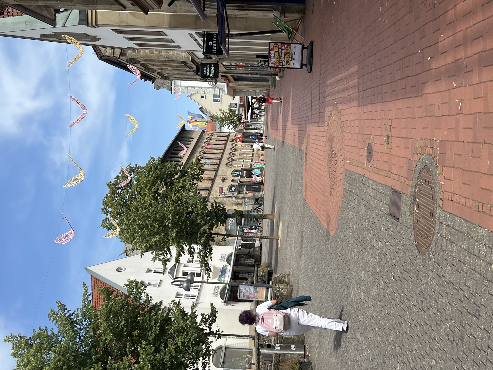 In the city of Unknown, Unknown, on a sunny day, a woman with purple hair, wearing a white t-shirt, white pants, and a light pink backpack, walks away from the camera down a cobblestone path. She carries a dark green jacket over her left shoulder. The path transitions to red brick further down the street. On the left side of the path, a white building with large arched windows and a gabled roof is visible, with green trees in front. On the right side of the path, a row of multi-story buildings with stone facades lines the street. Decorative yellow and pink butterfly-shaped ornaments are strung across the sky above the buildings. Several shops are indicated by signage, including "Bücher," "Thalia," "Bredy," "Stadt-Parfumerie Pieper," and "famano." A large sign advertising a "MEGA PREISSTURZ" with discounts up to 70% is positioned on the right side of the path. Other pedestrians are visible in the distance, and a small white dog is present on the right. Bicycles are parked near some of the buildings. The sky is blue with scattered white clouds.