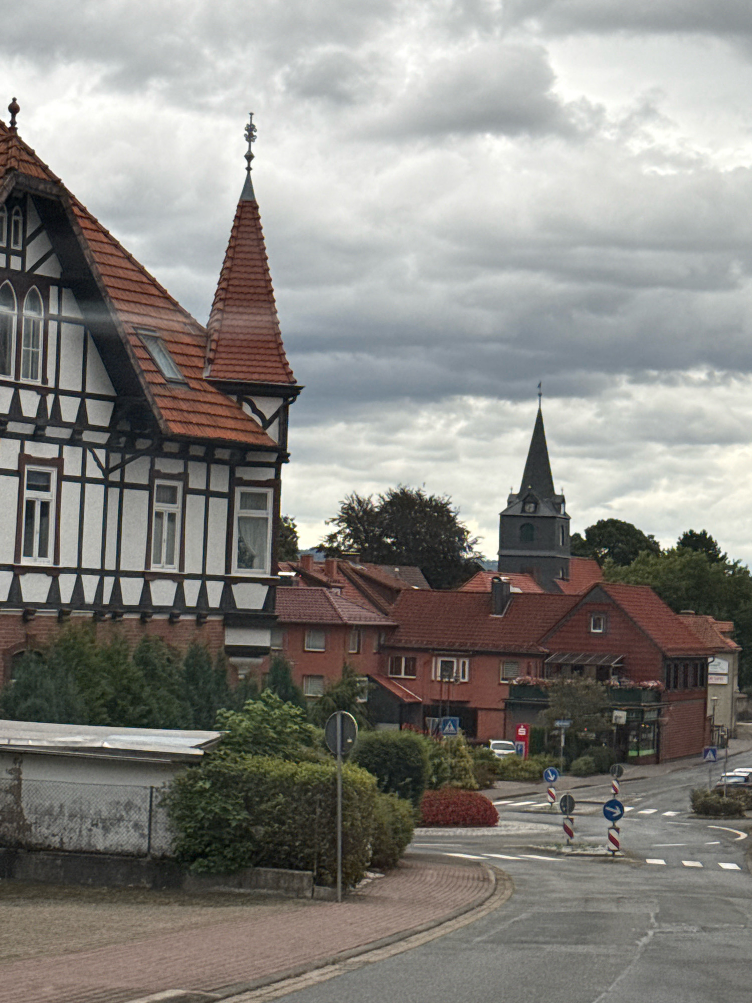 The image shows a street scene in Bad Sachsa, Germany, on an overcast day. In the foreground, a paved street curves to the left, bordered by a brick sidewalk. To the left of the street is a light-colored building with a fenced enclosure and green bushes. Further back, a traditional half-timbered house with a red tiled roof is visible. To the right of the half-timbered house, several red-tiled buildings form a small commercial area, with shops and awnings. In the background, a church steeple with a clock tower rises above the rooftops. The sky is filled with heavy, grey clouds. Traffic signs and road markings are present, indicating an intersection. No people are visible in the scene.