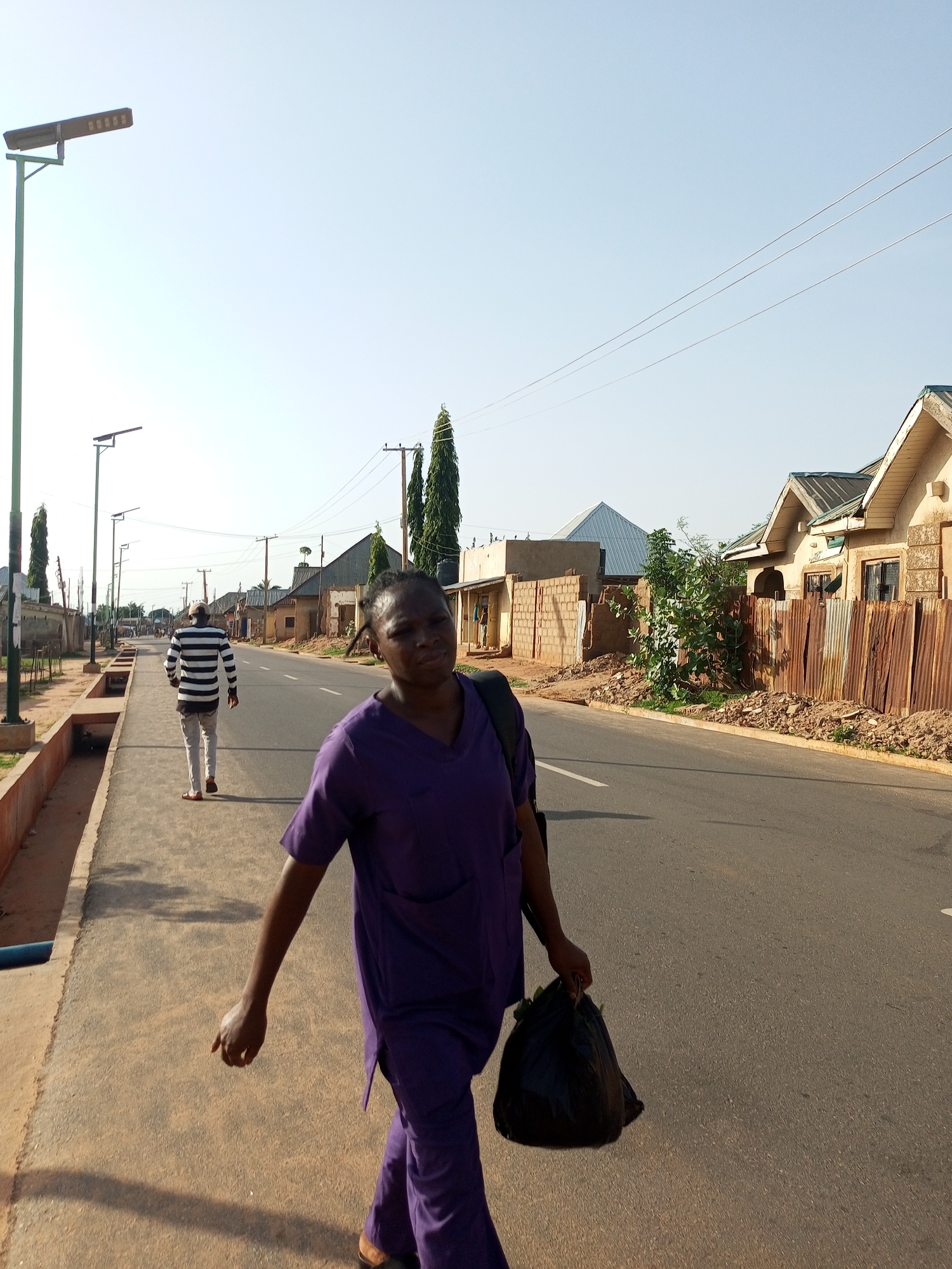 A woman in purple scrubs walks down a paved road in Tempure, Nigeria, carrying a black plastic bag in her right hand. Her face is turned towards the camera, and she appears to be looking at the viewer with a neutral expression. Behind her, a man in a black and white striped shirt and light-colored pants walks away from the camera, also on the paved road. The road is lined with buildings, some of which appear to be houses or small shops, with corrugated metal fences in front of some. There are streetlights along the side of the road, and utility poles with wires are also visible. The sky is clear and blue, suggesting it is daytime, likely morning or afternoon given the shadows. The scene appears to be a typical street in a residential or mixed-use area of Tempure.