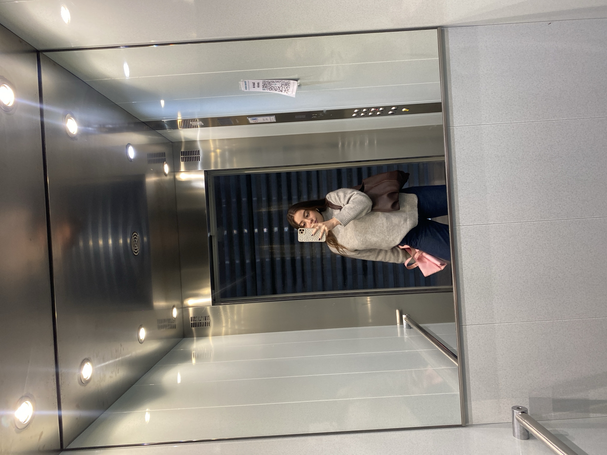 The image depicts a reflection of a young woman taking a selfie in an elevator. The elevator has a mirrored wall and is lined with stainless steel and white panels. There are round recessed lights in the ceiling and wall of the elevator. The reflection shows a glimpse of the elevator's control panel, a notice, and the closed doors behind the subject. The woman is wearing a light-colored sweater and dark pants. She is holding a phone with a patterned case. There is a brown backpack slung over her shoulder, and she is carrying a pink tote bag. The location is an elevator in the city of Unknown, Unknown.
