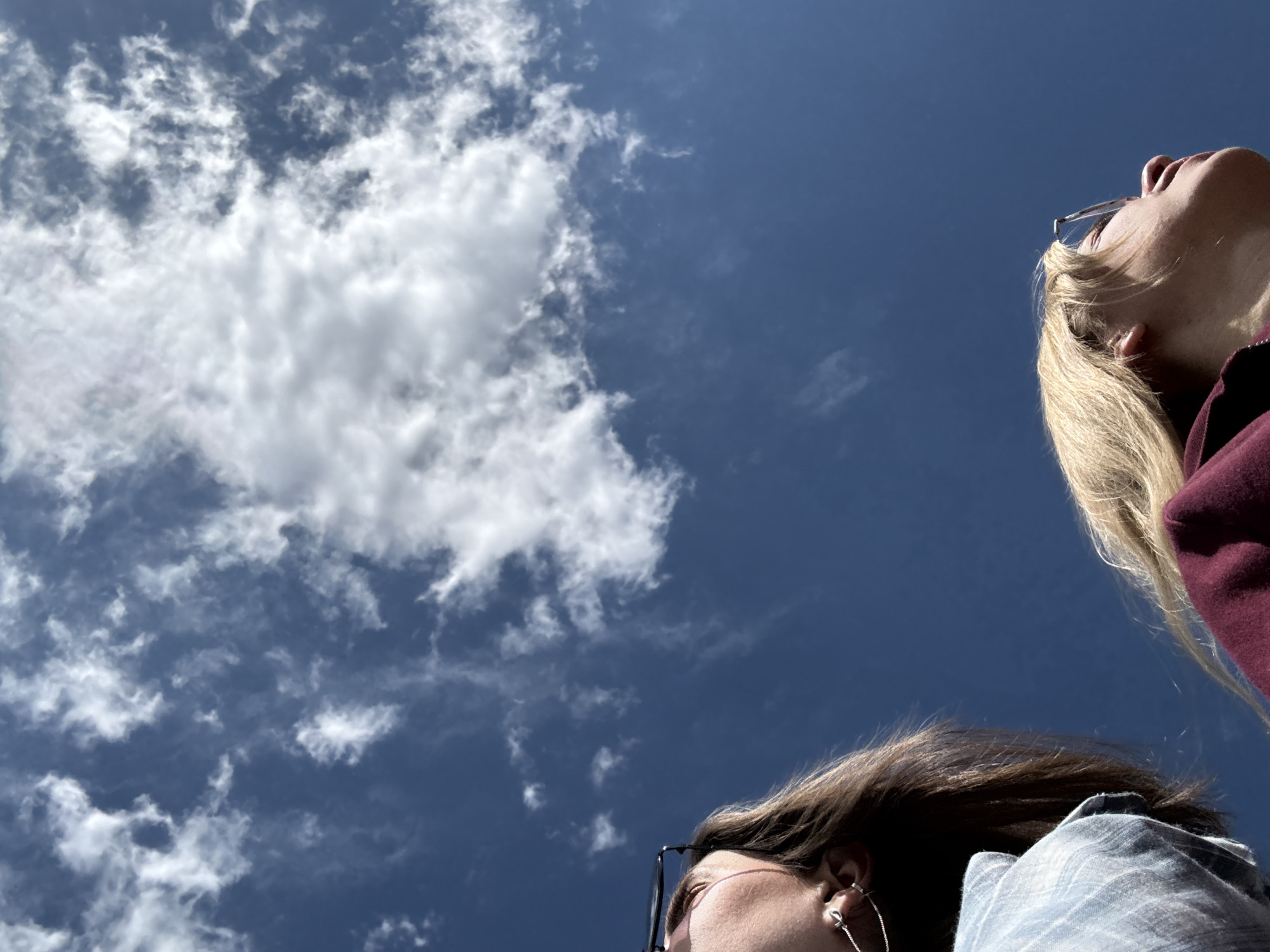 Two people are looking up at a sky filled with scattered clouds against a dark blue background. The person on the right has blonde hair and is wearing a maroon shirt. The person on the left has brown hair, is wearing glasses, and a light blue shirt. It is not possible to definitively determine the location is Krasnoyarsk, Russia based on the visual information provided.
