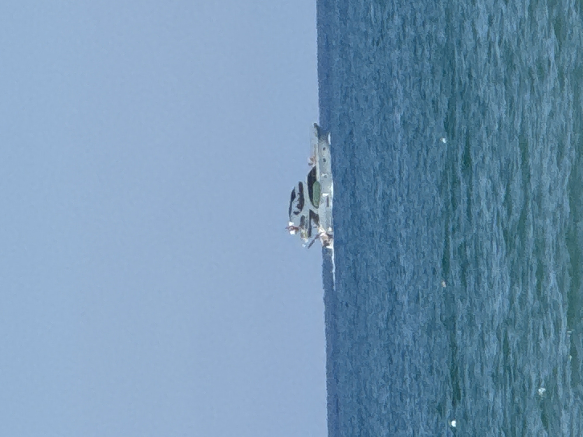 An aerial view of a yacht sailing on the sea off the coast of Manavgat, Turkey. The yacht is white and appears to be a luxury model. It is sailing in a calm, blue sea under a clear sky. The horizon is visible in the distance, separating the sea from the sky. The image captures a serene moment of a vessel cruising through the water.