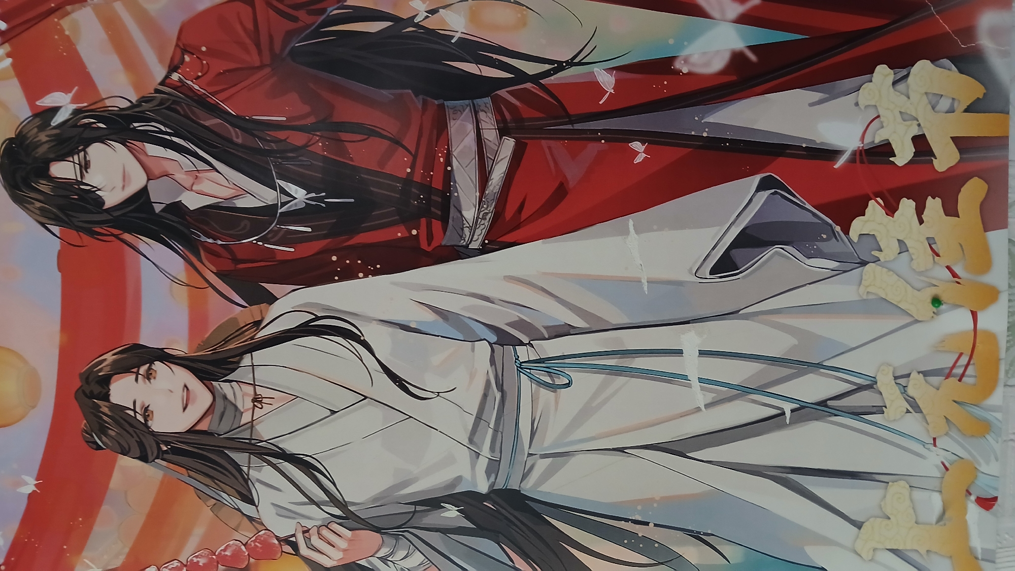 The image shows two cartoon characters, one in red and one in white, lying side by side. The figure in red is positioned above the figure in white. The background features indistinct shapes and colors, and there are golden, stylized characters or decorations on the right side of the image. The location is Unknown, Unknown.