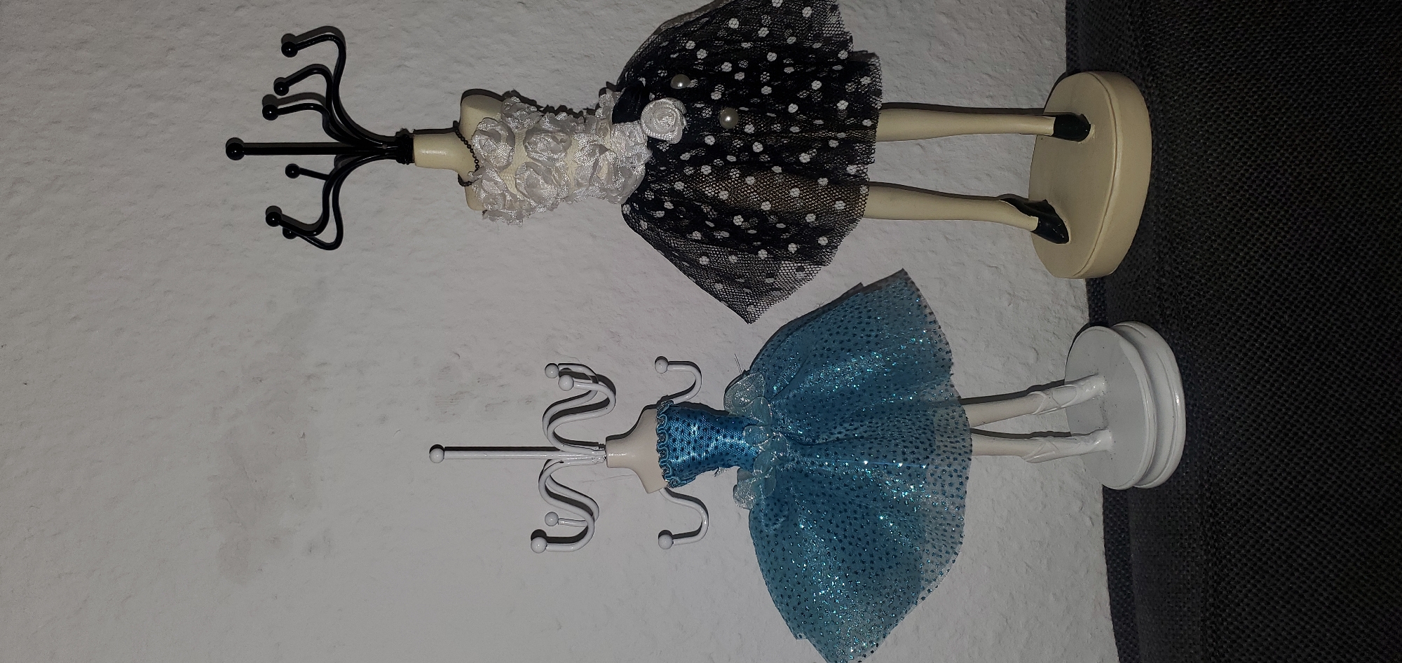The image displays two decorative mannequins designed to hold jewelry, set against a textured white wall. The mannequin on the left is adorned with a sparkling blue tutu-style dress and stands on a white circular base with ballet slippers. A white jewelry stand, shaped like a rack with multiple hooks, is positioned above its torso. The mannequin on the right wears a black and white polka dot skirt and a white ruffled top, accessorized with a dark necklace. It stands on a beige oval base and is topped with a black jewelry stand featuring antler-like hooks. Both mannequins have visible legs, with the one on the right wearing black wedge heels. The setting appears to be indoors, possibly on a dark fabric surface in what could be a residential environment in Sasbach, Germany.