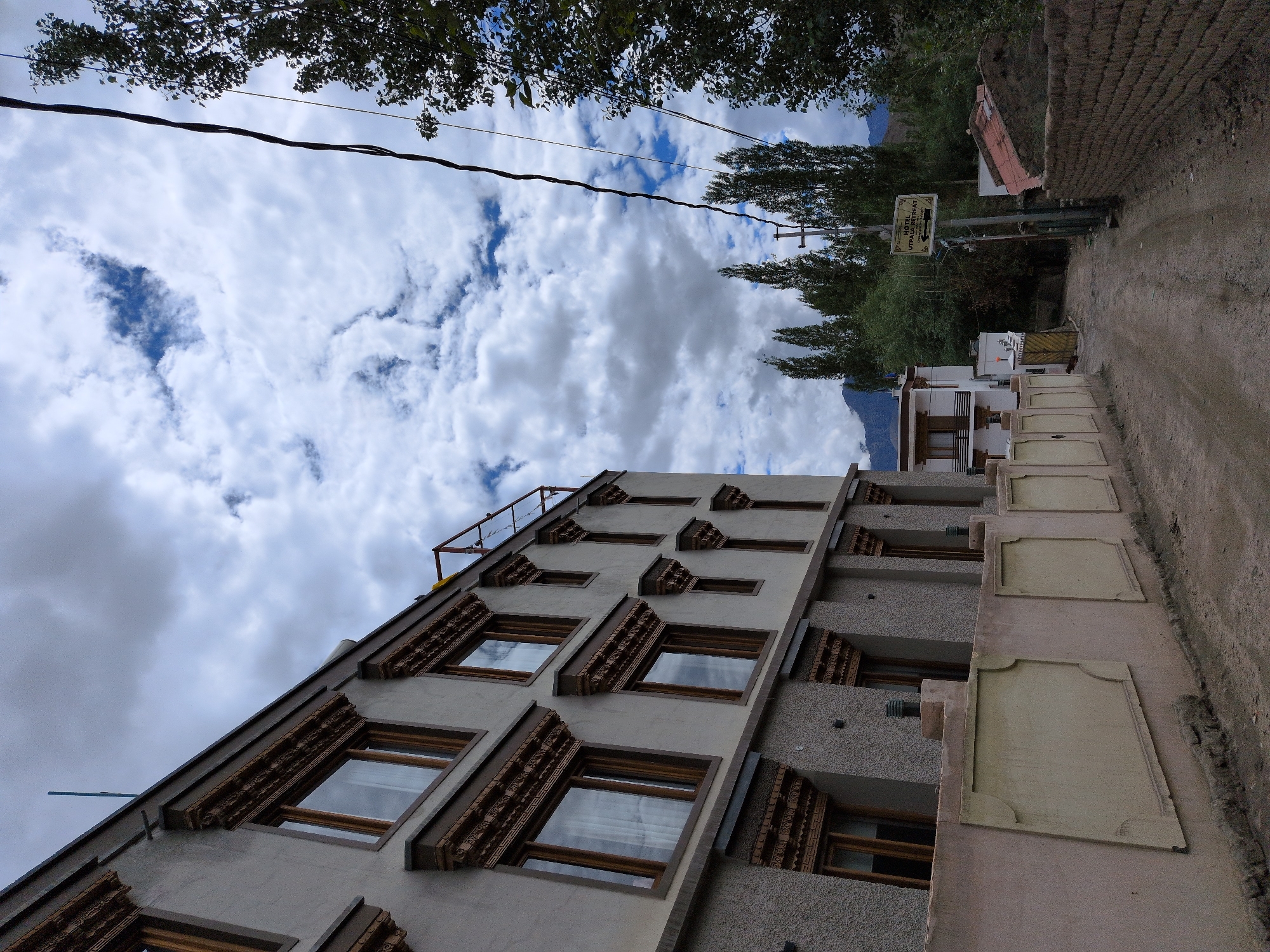 The image depicts a street scene in Leh, India. On the left, a multi-story building with traditional Tibetan architectural elements, such as decorative wooden window frames and ornate cornices, dominates the foreground. The building's facade is a pale beige color, with textured grey plaster on the lower levels. A low, decorative wall runs along the street in front of the building, featuring inset rectangular panels.
To the right of the street, trees and a brick wall are visible. In the background, a dirt road winds through the scene, leading towards a cluster of buildings and lush green trees. A signpost with an arrow and text reading "HOTEL UTPAAL RETREAT" is situated on the right side of the road, indicating the presence of accommodations. Mountains are visible in the far distance, partially obscured by trees and clouds. The sky is overcast with dramatic cloud formations. The overall impression is one of a peaceful, somewhat rustic environment in the Himalayan region. There are no people visible in the image.
