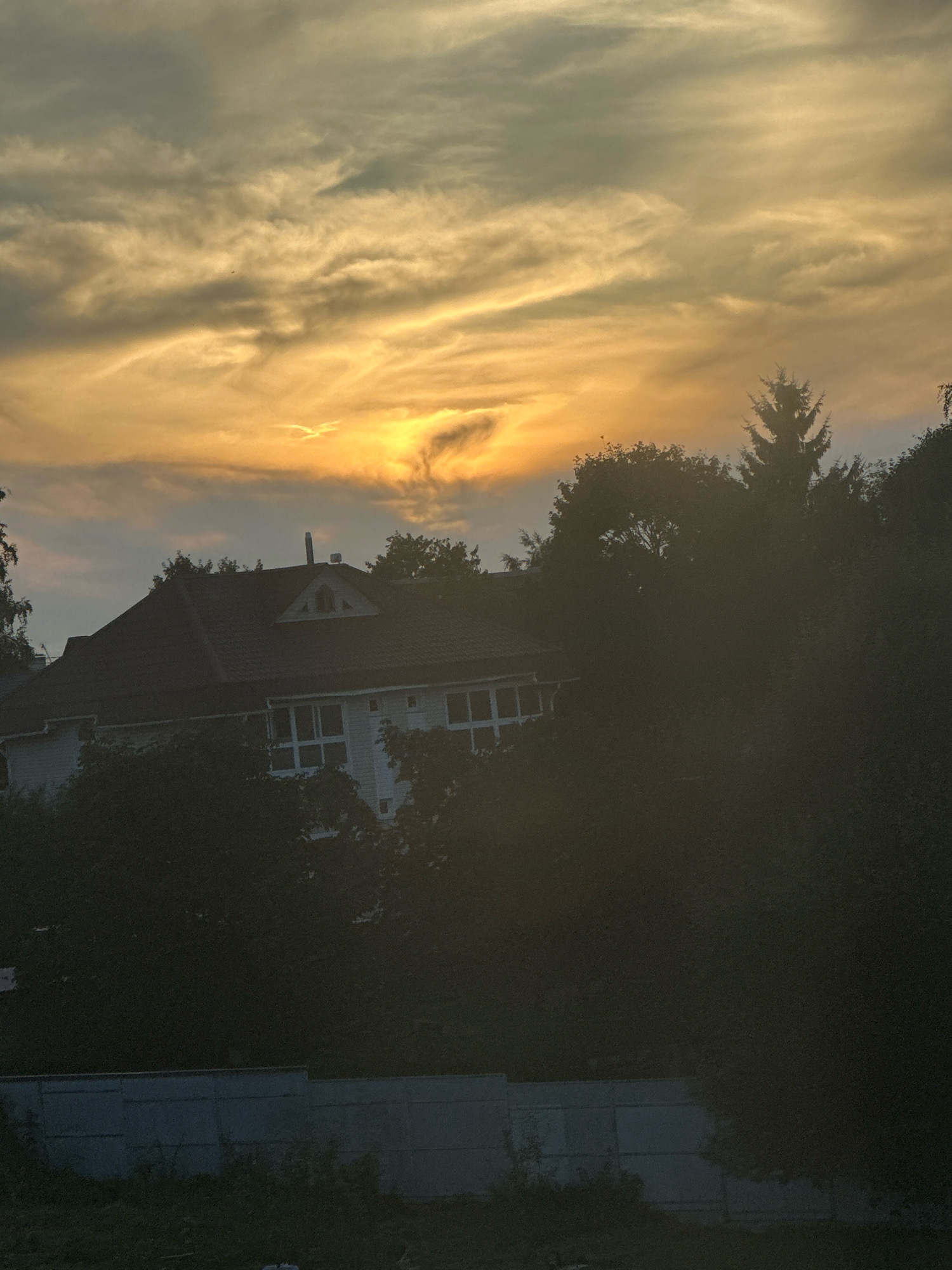 The image captures a dusky sky over a residential area, possibly in Virki, Russia. The sky is filled with wispy clouds, illuminated by the warm, orange glow of a setting sun. A white house with a dark roof is visible through a dense arrangement of trees and foliage in the foreground. The house features a prominent gable with a small window. A grey concrete fence runs along the bottom of the frame, partially obscured by vegetation. The scene is otherwise devoid of people or any discernible activity, presenting a quiet, twilight atmosphere.
