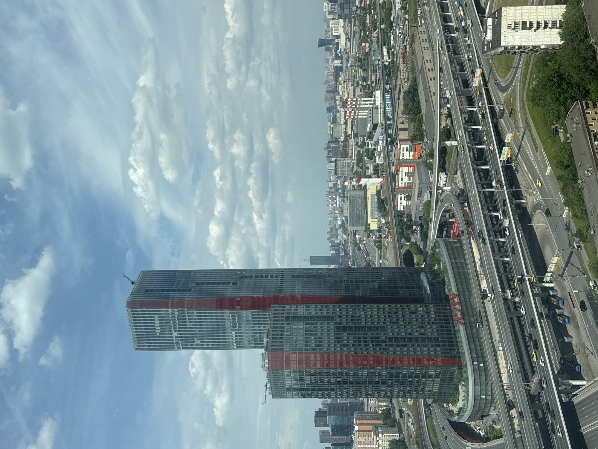 This is an aerial, eye-level, wide-angle photograph of the city of Khimki, Russia, under a partly cloudy sky. The foreground is dominated by a multi-layered highway interchange, with numerous vehicles traversing the various levels. The buildings in the image include two prominent, modern skyscrapers with dark facades and red vertical accents. The taller skyscraper is topped with a construction crane. To the right of the skyscrapers, the cityscape extends into the distance, featuring a mix of industrial buildings, smaller residential structures, and distant skyscrapers, one of which has a distinctive curved silhouette. The overall environment appears to be a dense urban area with significant infrastructure, including roads and industrial facilities.