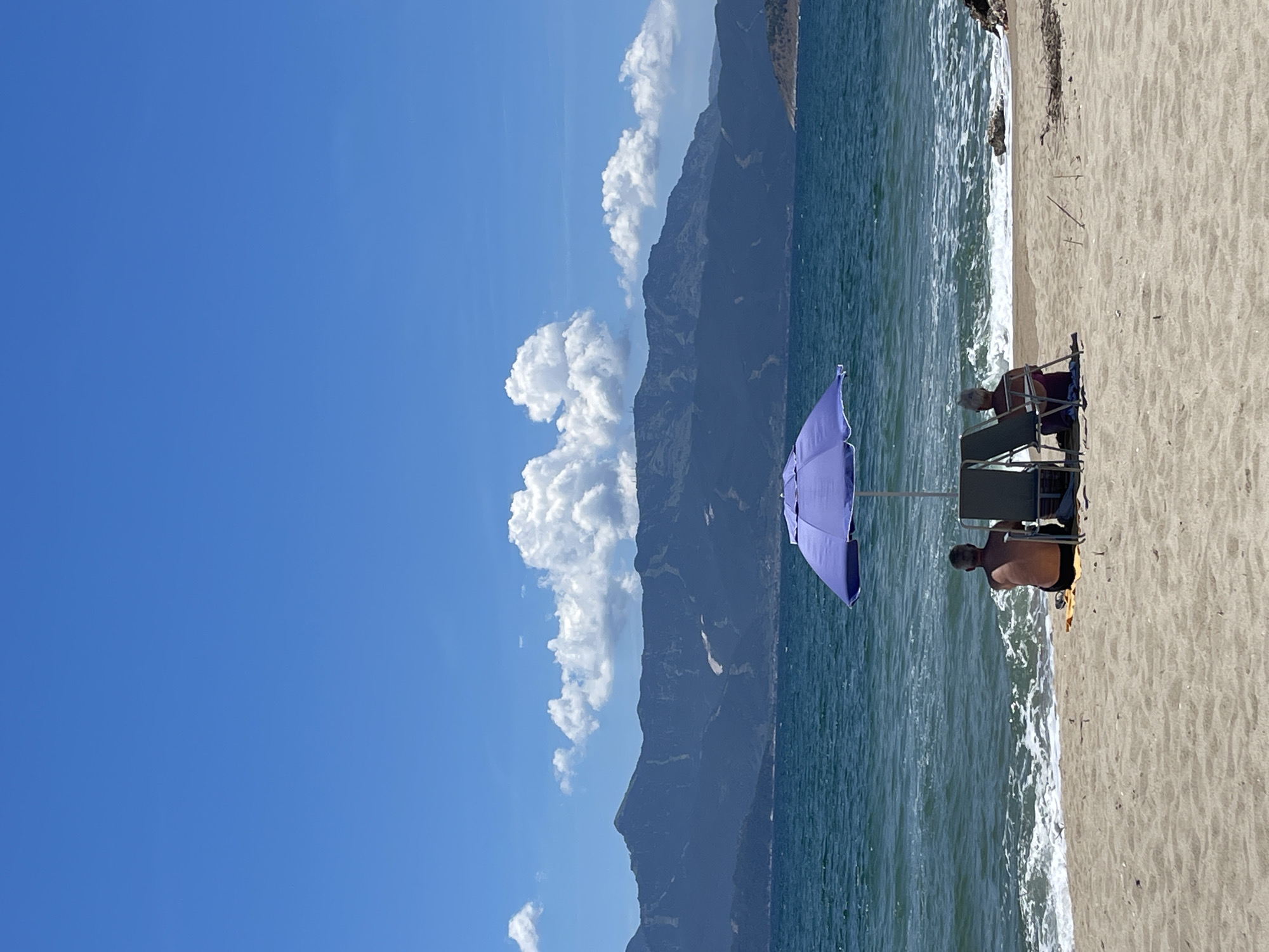 This image captures a serene beach scene in Keramoti, Greece. In the foreground, a sandy beach stretches towards the clear blue waters of the sea. Two individuals are seated on beach chairs under a purple umbrella, facing the water, seemingly enjoying the view. The sea is calm with gentle waves lapping at the shore. In the background, a majestic mountain range forms a dramatic backdrop against a bright blue sky dotted with fluffy white clouds. The overall atmosphere is one of peaceful relaxation on a beautiful summer day.