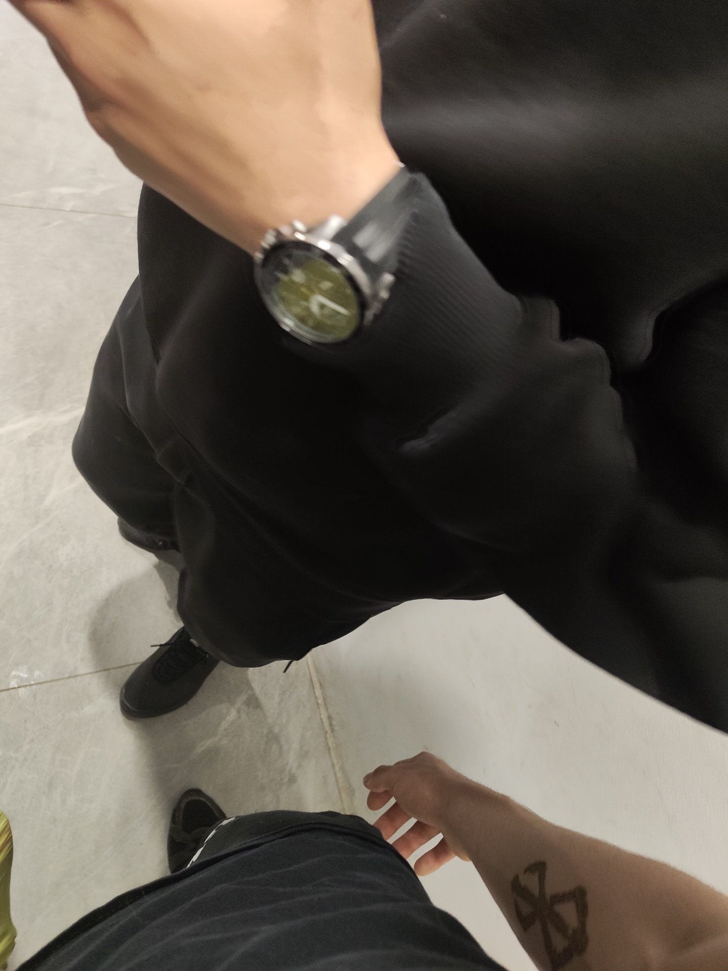 A top-down view shows a person wearing a black hoodie and black pants, standing on a light-colored tiled floor. The person has a watch on their left wrist. In the bottom right corner, another person's arm is visible, with a tattoo on their forearm. Their black pants and shoes are also visible. The location is unknown.