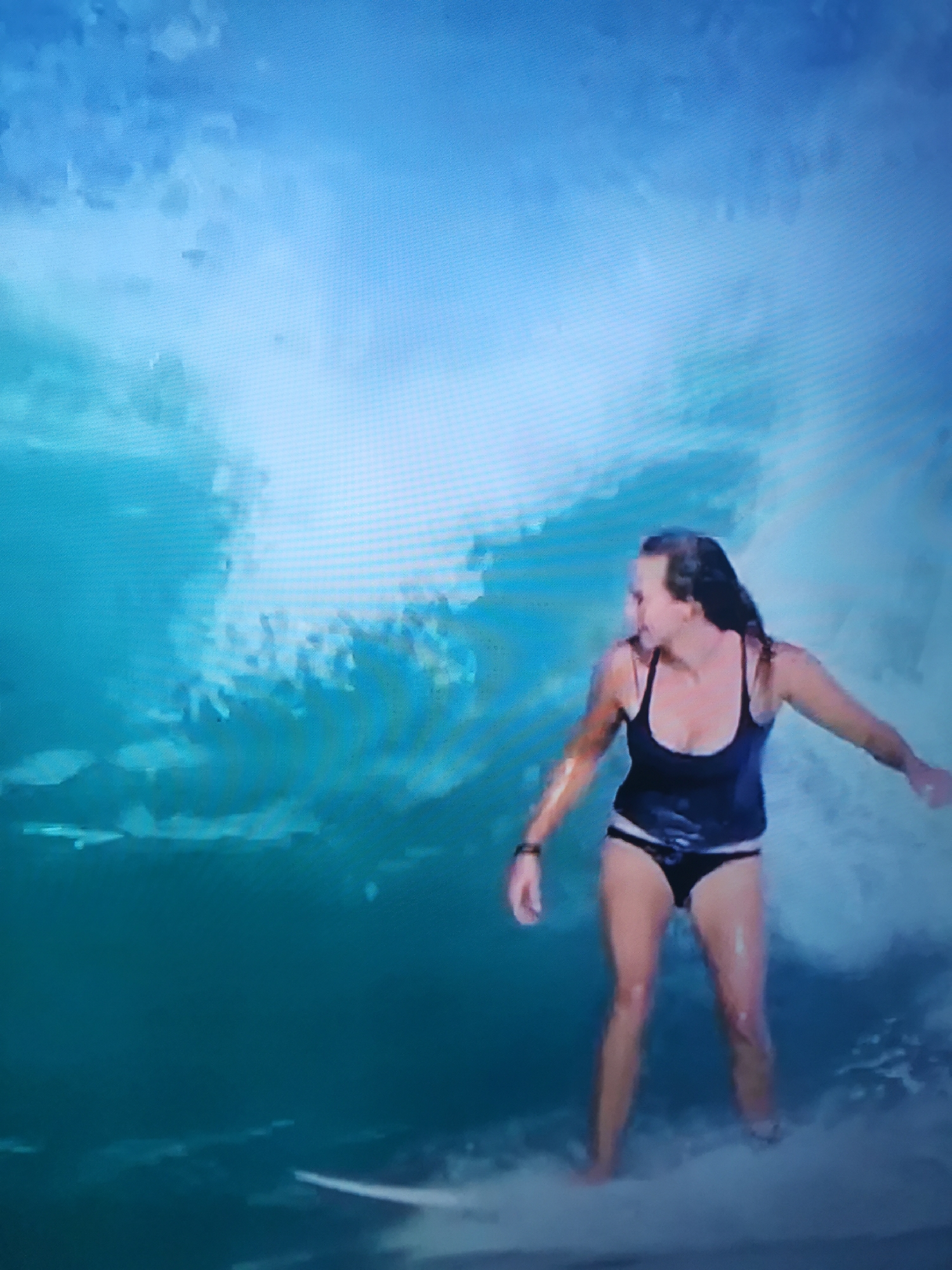 This media file depicts a young woman surfing on a wave. She is wearing a black swimsuit and has her hair pulled back. The setting is a body of water with a large wave cresting behind her, creating a blue and white environment. The woman appears focused and engaged in the activity. The image has a slightly grainy quality, suggesting it might be a capture from a video or a photograph taken in low light conditions. There is no visible text within the frame.
