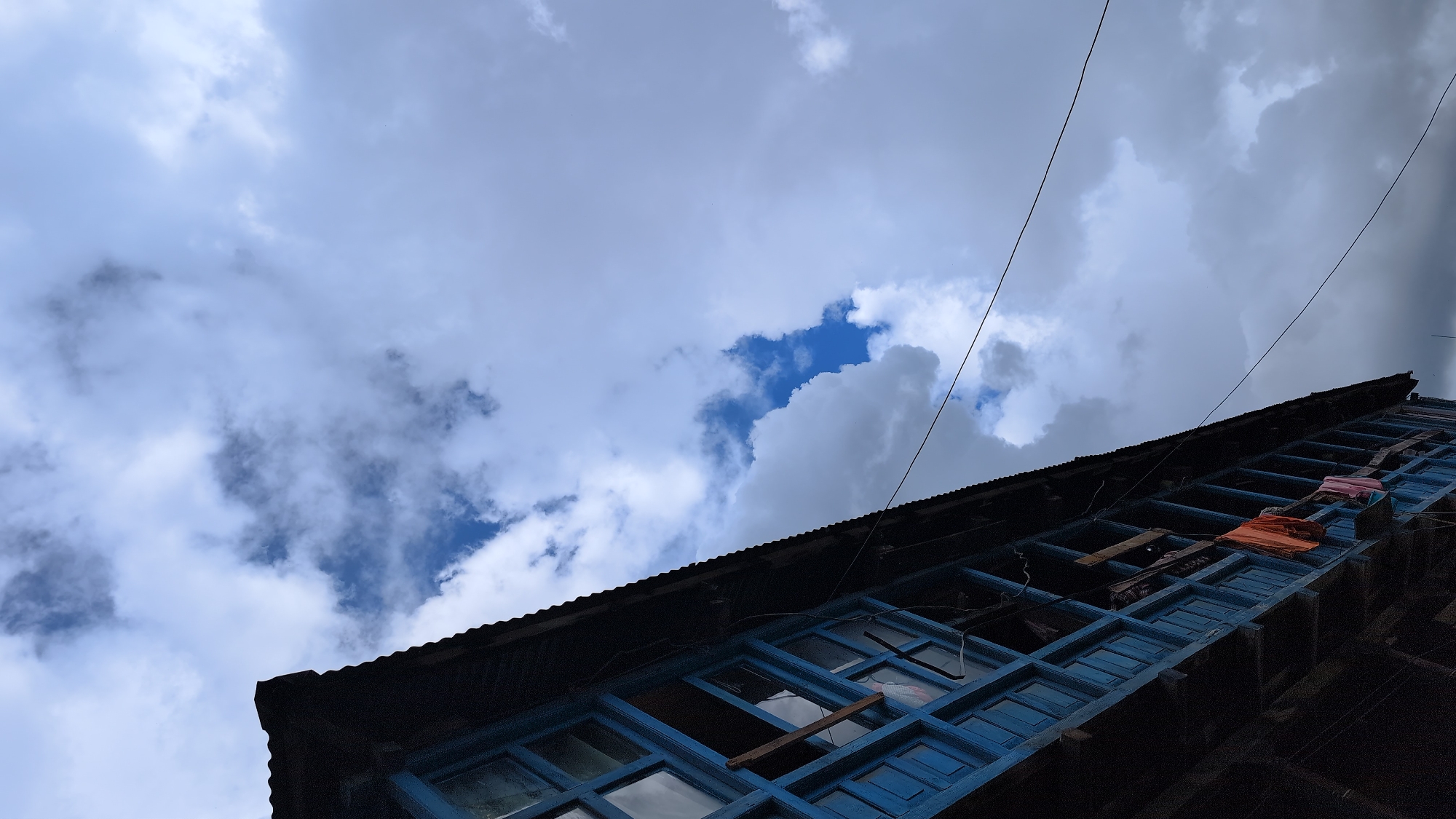 The image shows the exterior of a traditional multi-story building with a dark wooden facade and bright blue painted window frames, possibly in Vashisht, India. Laundry, including an orange and pink garment, is hanging from the balconies. The building is set against a dramatic sky filled with heavy, grey and white clouds, with patches of bright blue sky visible. Two thin dark wires are stretched across the sky. The perspective is from below, looking upwards at an angle.
