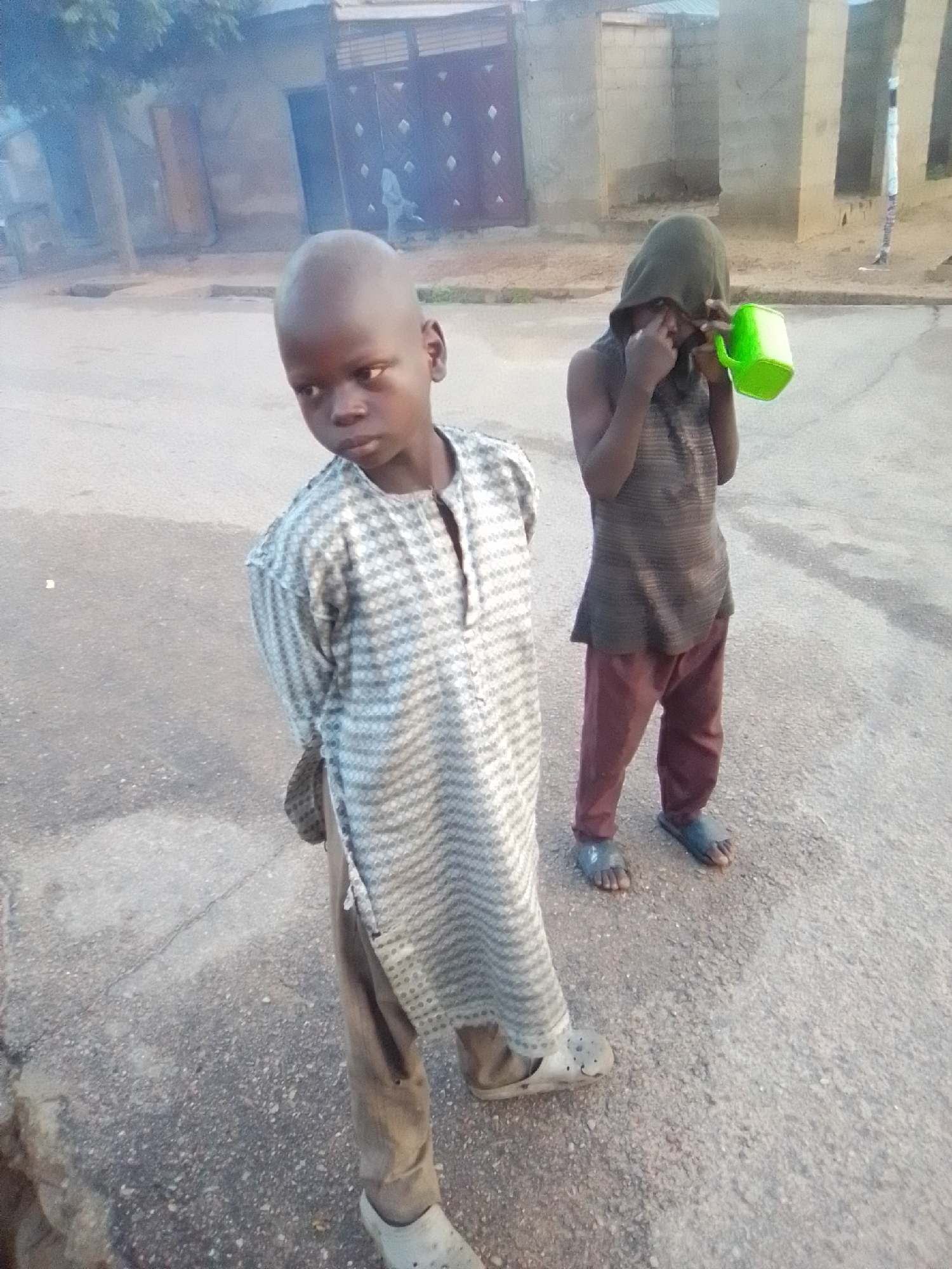 In Liji Hill, Nigeria, two young boys stand on a paved street. The boy on the left, with a shaved head, wears a patterned tunic and brown trousers. The boy on the right, with a dark green hood pulled over his head, wears a dark sleeveless shirt and maroon trousers. He holds a green plastic cup in his right hand and appears to be wiping his eye with his left. Behind them, a building with a textured wall and barred windows is visible, along with a tree and a partial view of a closed gate. The ground is a mix of asphalt and loose gravel.