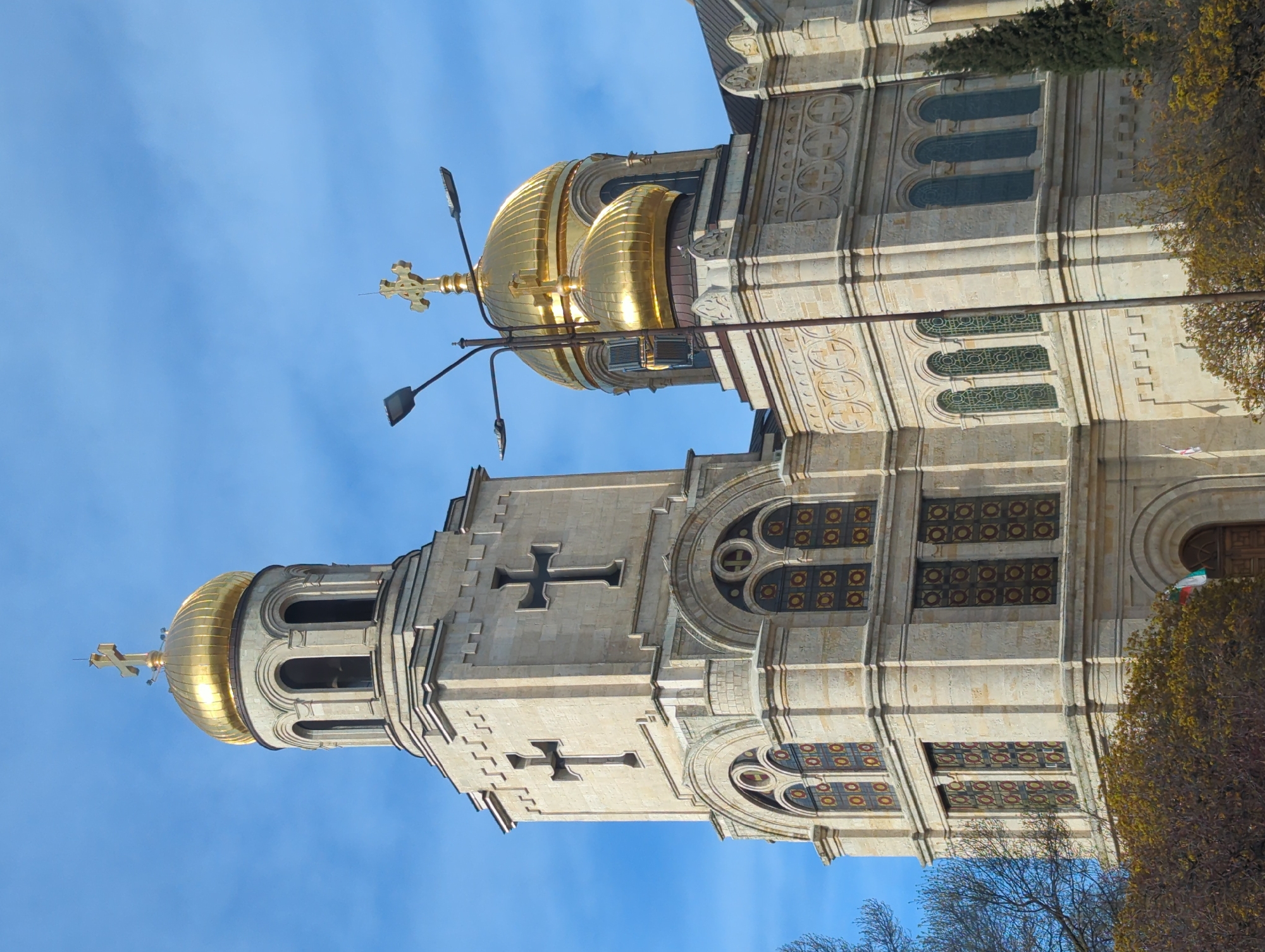 The image captures the Assumption Cathedral in Varna, Bulgaria. It features two prominent structures: a bell tower with a golden dome and a larger cathedral building with a golden dome. Both structures are light-colored stone. The tower has large crosses on its facade and windows with dark mullions. The cathedral building has patterned windows and detailed ornamentation. The sky is blue with a few clouds. Part of a street light is visible in the foreground, adding to the urban setting. Trees are partially visible on the right and bottom.