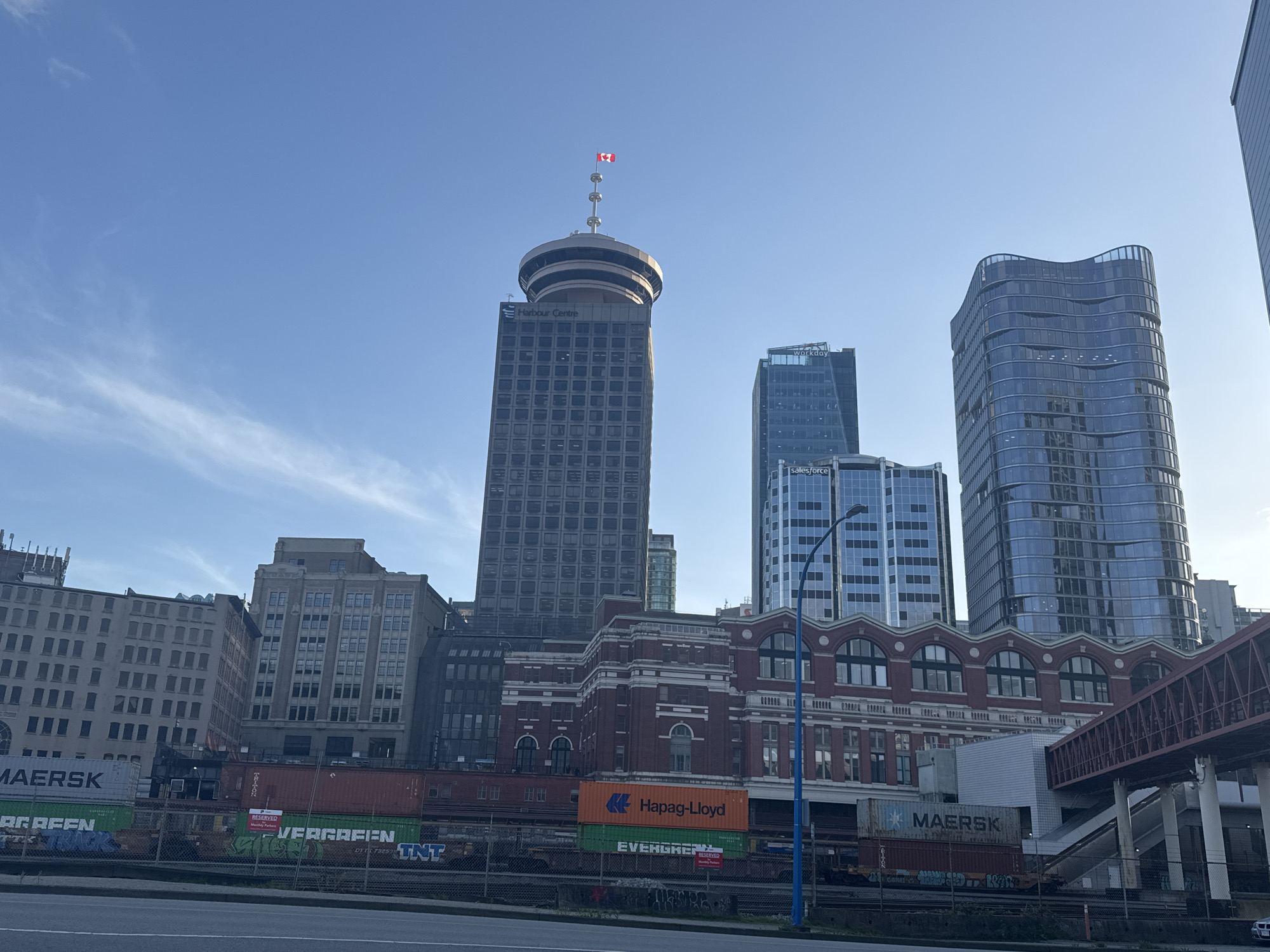 The image shows a cityscape of Vancouver, Canada, with a clear blue sky overhead. Several buildings are visible, including the Harbour Centre tower with a Canadian flag atop its spire, a glass-faced building displaying "workday salesforce", and a modern, curving building. In the foreground, a railway line is visible with freight containers labelled "MAERSK", "Hapag-Lloyd", and "EVERGREEN", some bearing graffiti. A historic red-brick building stands adjacent to the railway tracks, and a pedestrian overpass is seen on the right side of the frame. The scene captures a blend of the city's modern architecture and industrial transportation.