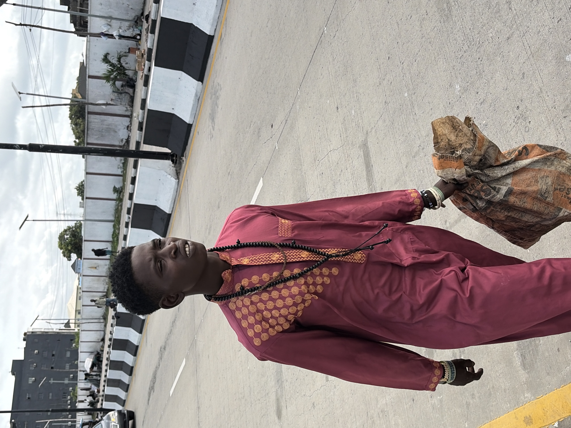A man stands on a road in Igboefon, Nigeria. He is wearing a maroon, long-sleeved traditional outfit with gold accents and a black beaded necklace. He is holding a large brown and orange patterned bag in his right hand. He has black hair and dark skin. The road has black and white striped curbs. In the background are buildings, streetlights, and other people. The sky is overcast.