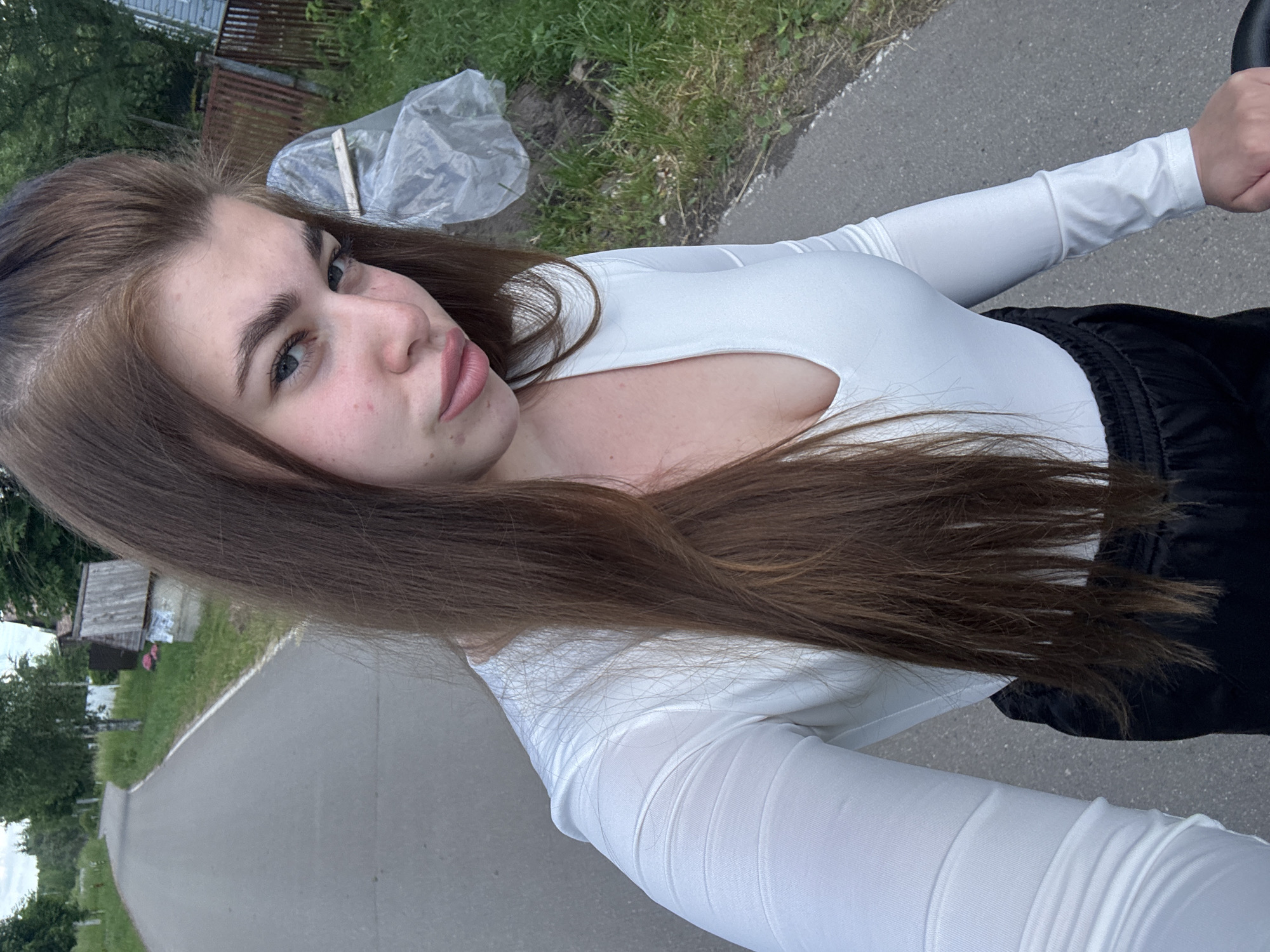 A young woman with long brown hair and fair skin, wearing a white long-sleeved shirt and black pants, is taking a selfie outdoors. She has a neutral expression, with her lips slightly pursed. The background suggests a rural or suburban setting, with a paved road curving to the left, bordered by grass and trees. In the distance, a small wooden structure and a fence are visible, along with what appears to be a covered object, possibly plastic sheeting. The lighting indicates it is daytime, likely late afternoon or early evening, with overcast skies. The photo appears to have been taken in or near Мещёры, Russia, based on the provided context.