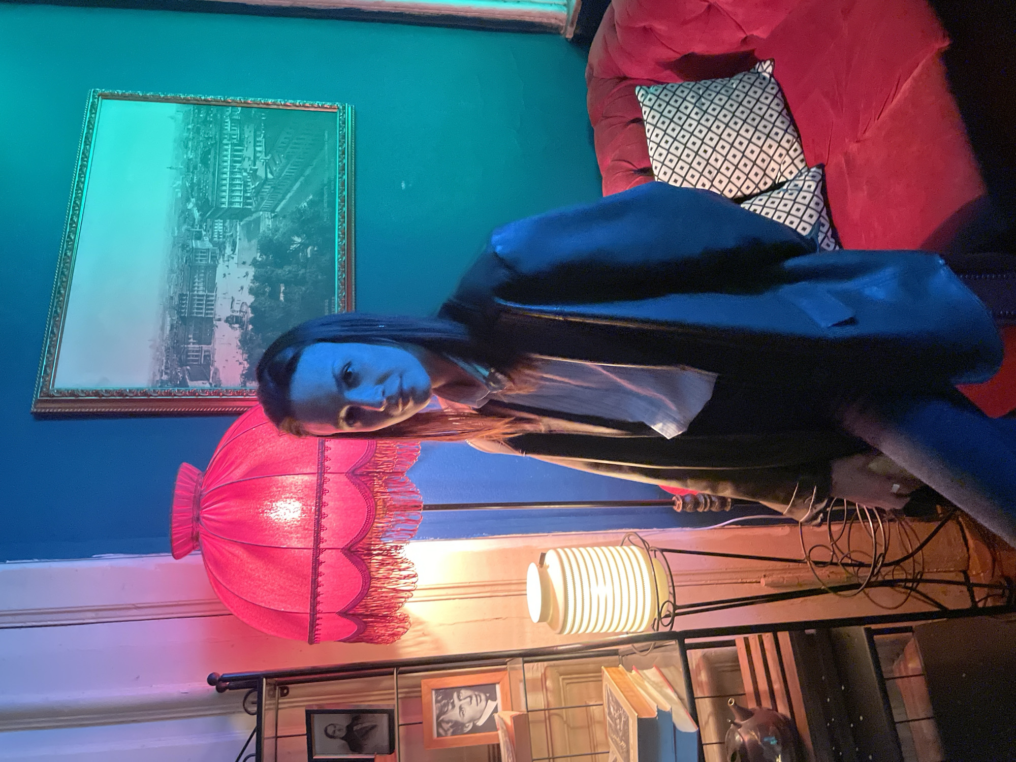 The image shows a person in a room with eclectic decor. The room features a blue wall adorned with a framed picture, and a red couch with a patterned pillow. A unique, fringed pink lamp stands near a shelf containing books, framed photos, and a decorative orb. Another lamp, cylindrical in shape, sits beside the pink one. The location is undetermined and there is no evidence suggesting it is in Saint Petersburg, Russia.