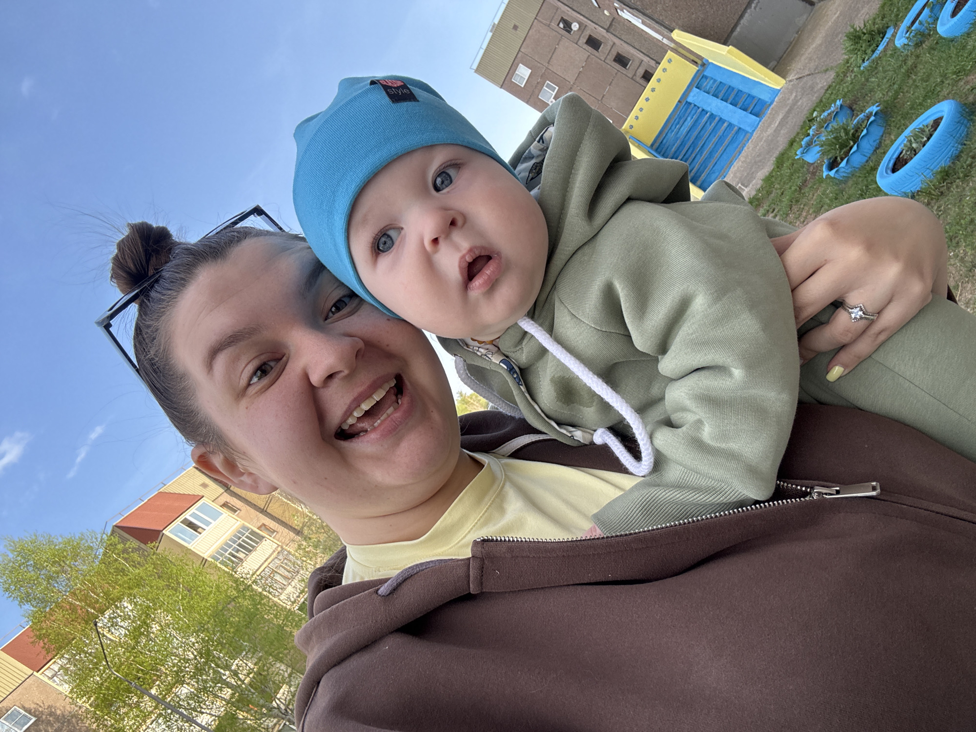 The image shows a woman holding a baby. The woman is wearing a brown hoodie over a yellow shirt and has glasses on her head, with her hair in a bun. She is smiling at the camera. The baby is wearing a light green hooded jumpsuit and a blue beanie. The background shows an urban environment, including a building and painted tires used as flowerbeds. The location is in the city of Марковский, Russia.