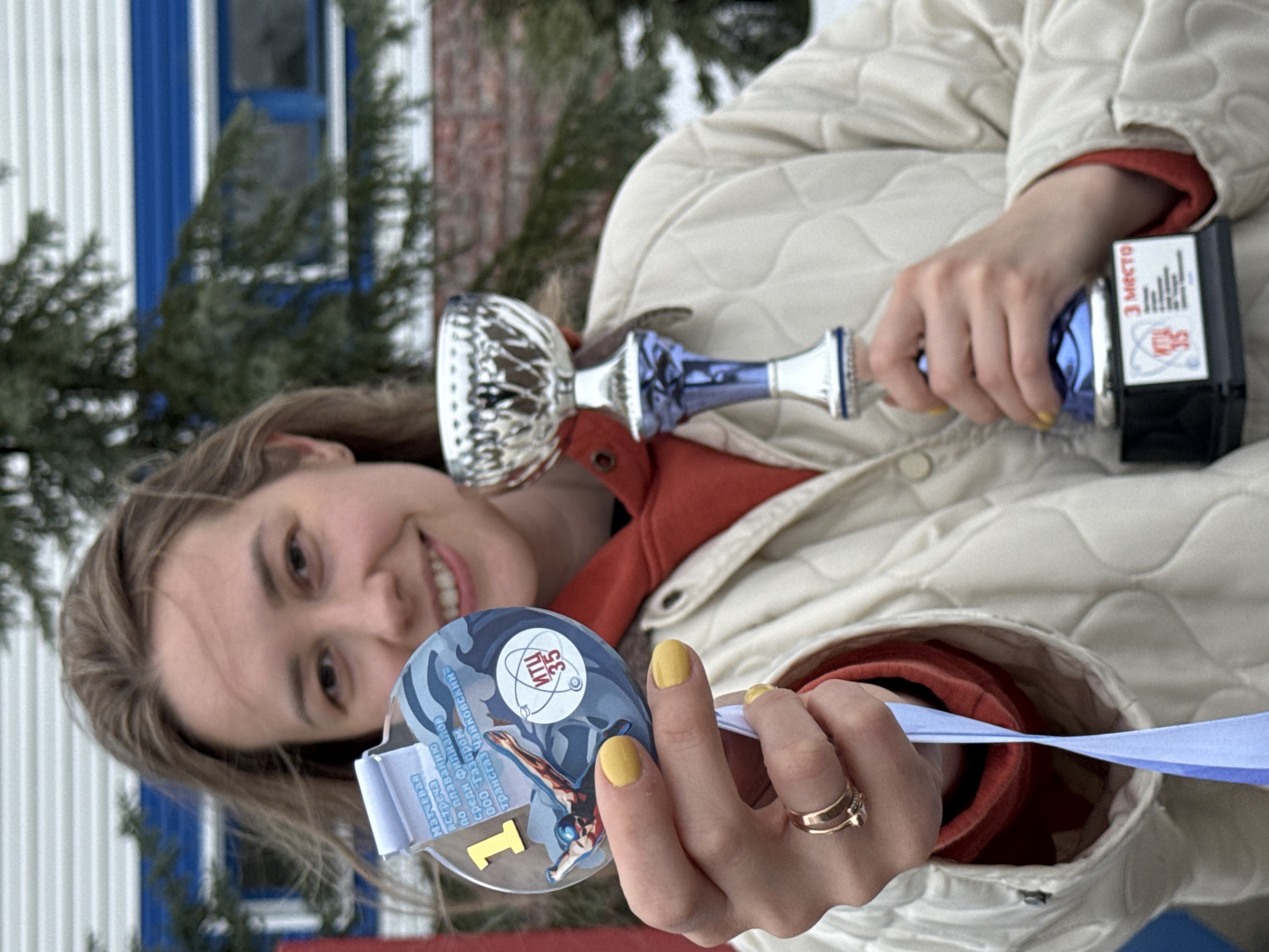 A woman with light brown hair smiles while holding a silver and blue trophy, a black trophy with a label that reads "3 место" (3rd place), and a medal with a blue ribbon. The medal shows a swimmer figure and the inscription "ИТЦ 35" (ITC 35) and "1" (one). Text on the medal is written in Russian; the text on the medal appears to refer to a swimming match among branches of Transgaz Chaikovsky. The woman wears a light tan quilted jacket over a red hoodie and a gold ring on her left ring finger. The background features a building with white siding and blue trim, as well as green foliage. The image suggests the location is Chaikovsky, Russia.