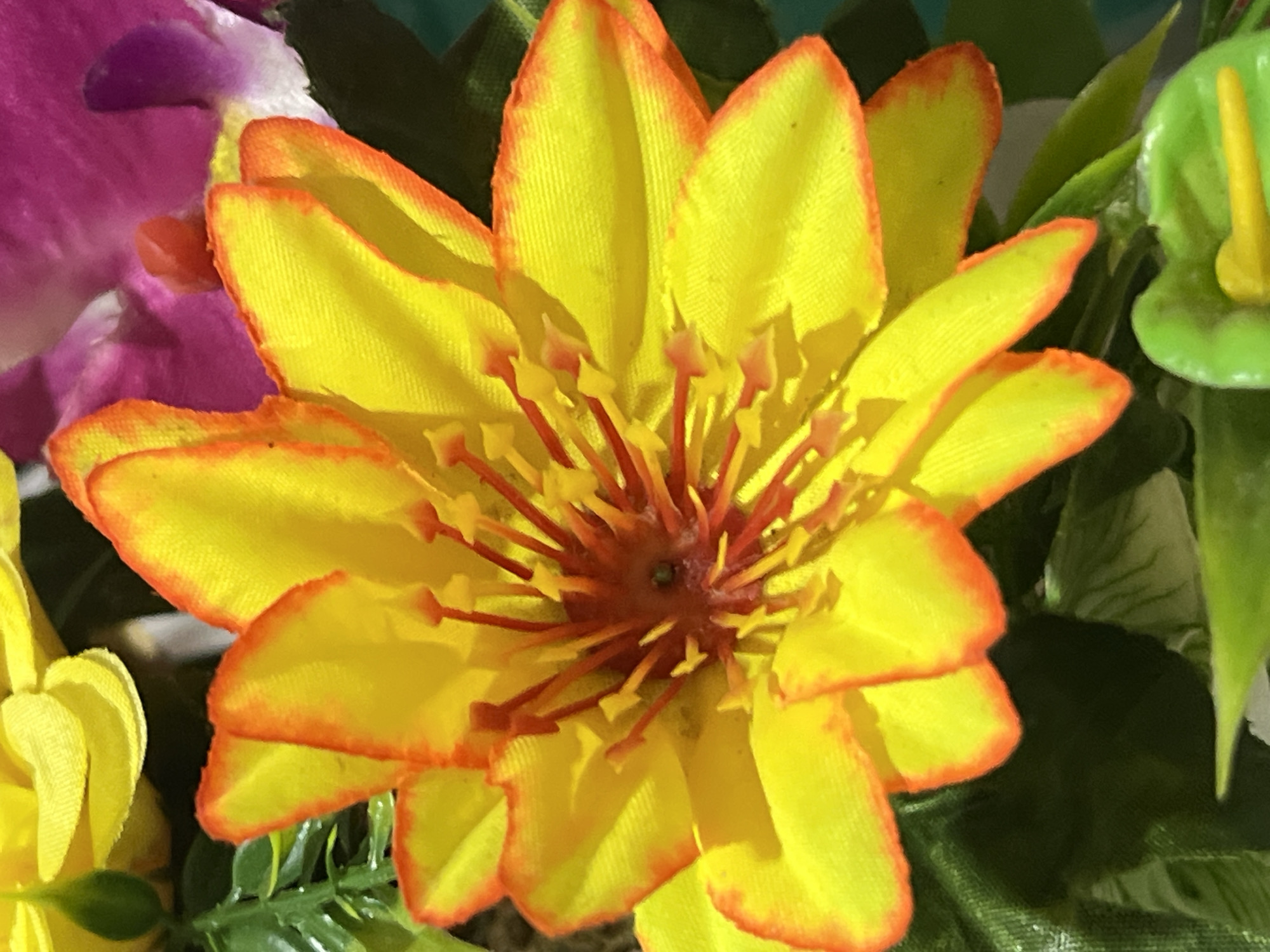 A close-up, overhead shot showcases a vibrant, artificial yellow and orange lotus flower in focus. The petals of the lotus are a bright yellow with fiery orange edges, and its center is adorned with numerous red and yellow stamen. Behind the lotus, parts of other artificial flowers are visible: a soft pink and white orchid on the right, and hints of yellow petals and green leaves on the left and bottom. The lighting suggests a bright, indoor setting, possibly a display or decoration. The image does not depict any specific location in Denpasar, Indonesia, nor does it contain any people or actions.