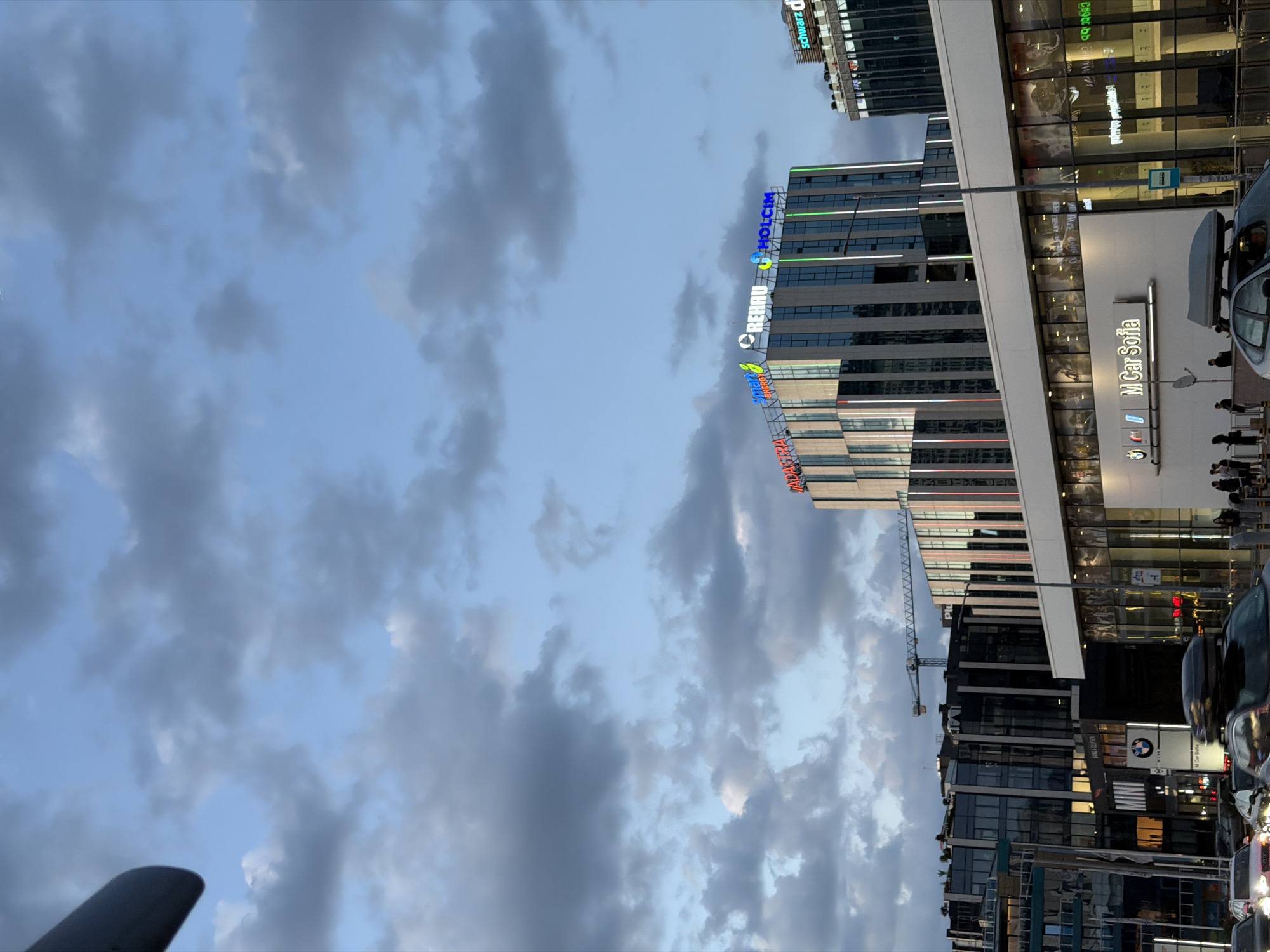 The image, rotated to a portrait orientation, captures an urban scene likely in Sofia, Bulgaria, evidenced by the "M Car Sofia" sign. The scene includes a mix of modern architecture, featuring buildings with glass facades and distinctive signage for companies like REHAU and HOLCIM. Construction is ongoing, suggested by the presence of a crane. The sky is cloudy, indicating either early morning or late evening. Cars and pedestrians are visible, suggesting an active urban environment. The angle and framing of the shot indicate it was taken from a street-level perspective.