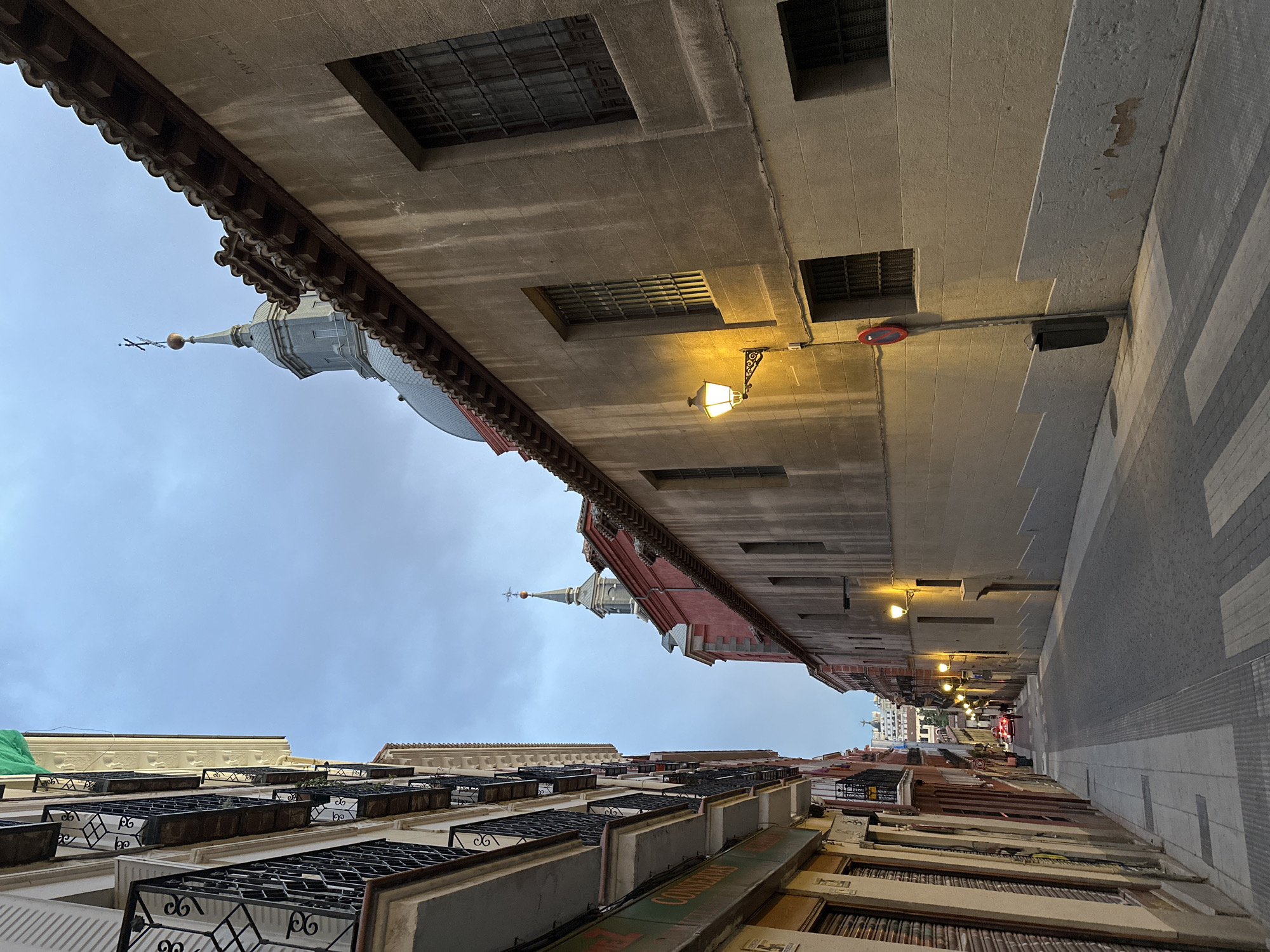 The image shows a narrow street in Madrid, Spain, viewed from a low angle, with buildings lining both sides. The perspective makes the buildings appear to lean inward and converge towards the sky, which is overcast with pale blue and grey clouds. The buildings have multiple floors with windows, some with decorative ironwork or grilles. Street lamps are visible along the buildings, casting a warm light. In the distance, there is a glimpse of a church steeple and other buildings further down the street. The street itself is paved and appears to be clean. There are no people visible in the image. Some of the buildings have signs and awnings visible.