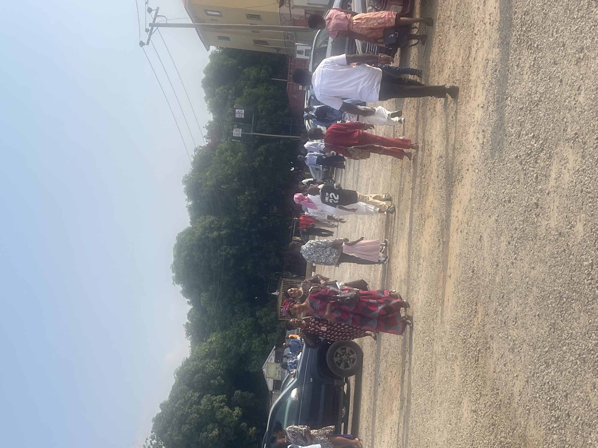 The image shows a gathering of people outdoors, likely in Abuja, Nigeria. The background includes trees, buildings, and utility poles. The ground appears to be a gravelly or dirt surface. A dark-colored SUV is visible. People are standing in small groups, some near the vehicle, and some dressed in formal wear. There is no clear action or event taking place, so they may be waiting or socializing. The sky is bright and light blue.
