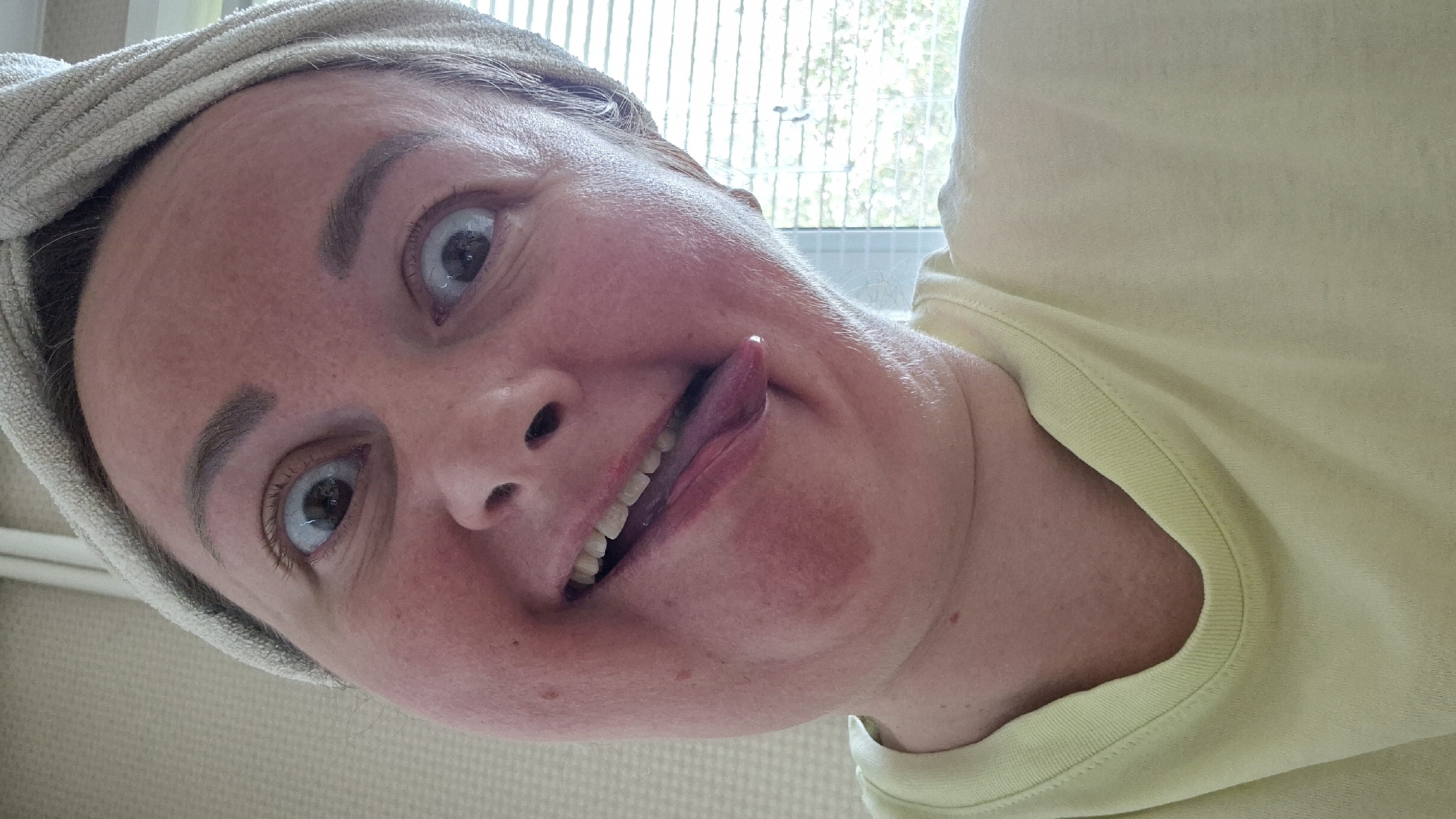 A close-up, selfie-style portrait captures a person with wide, surprised eyes and their tongue sticking out. They are wearing a light yellow t-shirt and a light-colored towel wrapped around their head. The person has fair skin with some redness on their cheeks and under their chin. The background appears to be an indoor setting with what looks like a window with blinds on the right side, allowing diffused daylight to enter. There is a textured wall visible on the left. The overall impression is one of playfulness or a candid, humorous moment. The image does not contain any elements that would allow for determination of location in Kemerovo, Russia.
