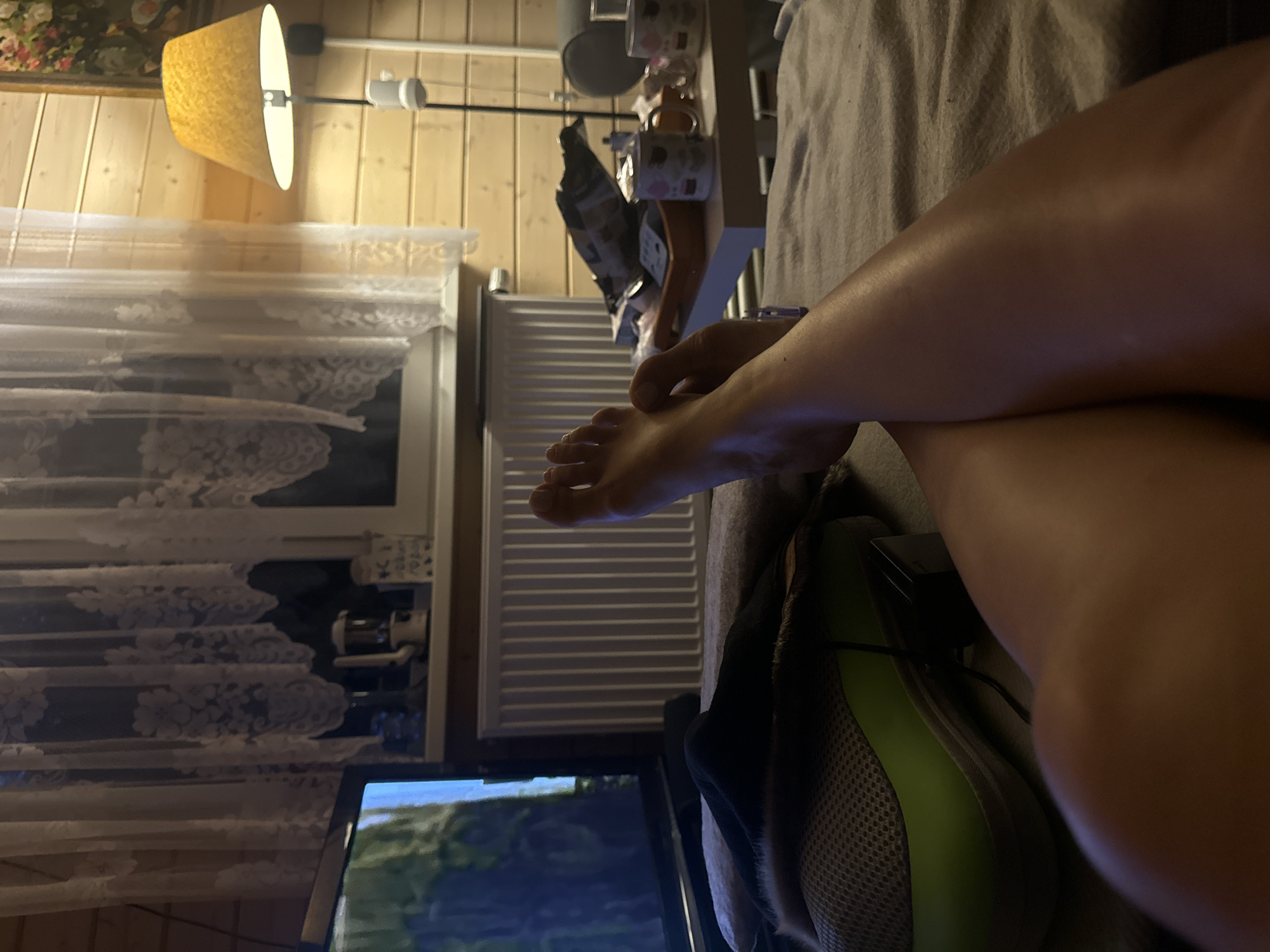 The image depicts a dimly lit indoor scene, likely a living room or bedroom, with wooden paneled walls. In the foreground, a person's legs and feet are crossed, resting on a bed or couch covered with a gray blanket. A television screen is visible on the left, showing a nature-related image. To the right, a side table holds several mugs, a tray with items, and a lamp with a yellow shade. A white radiator is positioned below a window with lace curtains. The overall impression is one of relaxation and leisure, with the television and comfortable seating suggesting a home environment. There is no discernible connection to the city of Stupino, Russia.