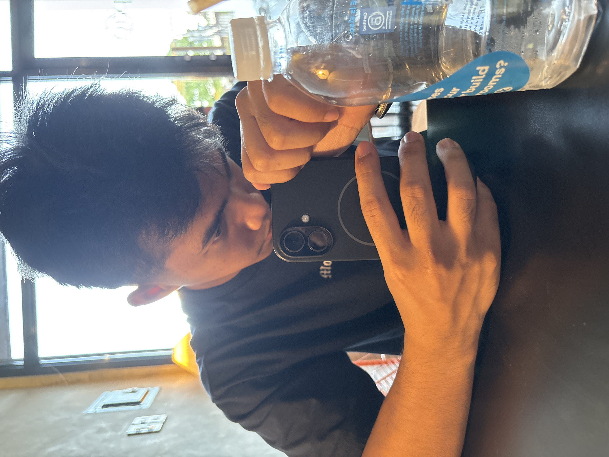 This is a close-up, angled shot of a person taking a selfie with a smartphone. The person is wearing a dark blue t-shirt and their dark hair is visible. They are holding a clear plastic water bottle with some liquid inside, and the bottle has a blue and white label with text. A smartphone is held in their hand, capturing the reflection of their face and the surrounding environment. The background shows a window with bright daylight streaming through, suggesting it is daytime. Outside the window, blurred green foliage is visible, indicating an outdoor or garden setting. The scene appears to be indoors, possibly at a cafe or restaurant, given the dark table surface. Some small white objects are on the table in the foreground. The text on the water bottle is partially visible and includes phrases like "build rooms?" and "Chat meet". The overall mood is casual and candid.