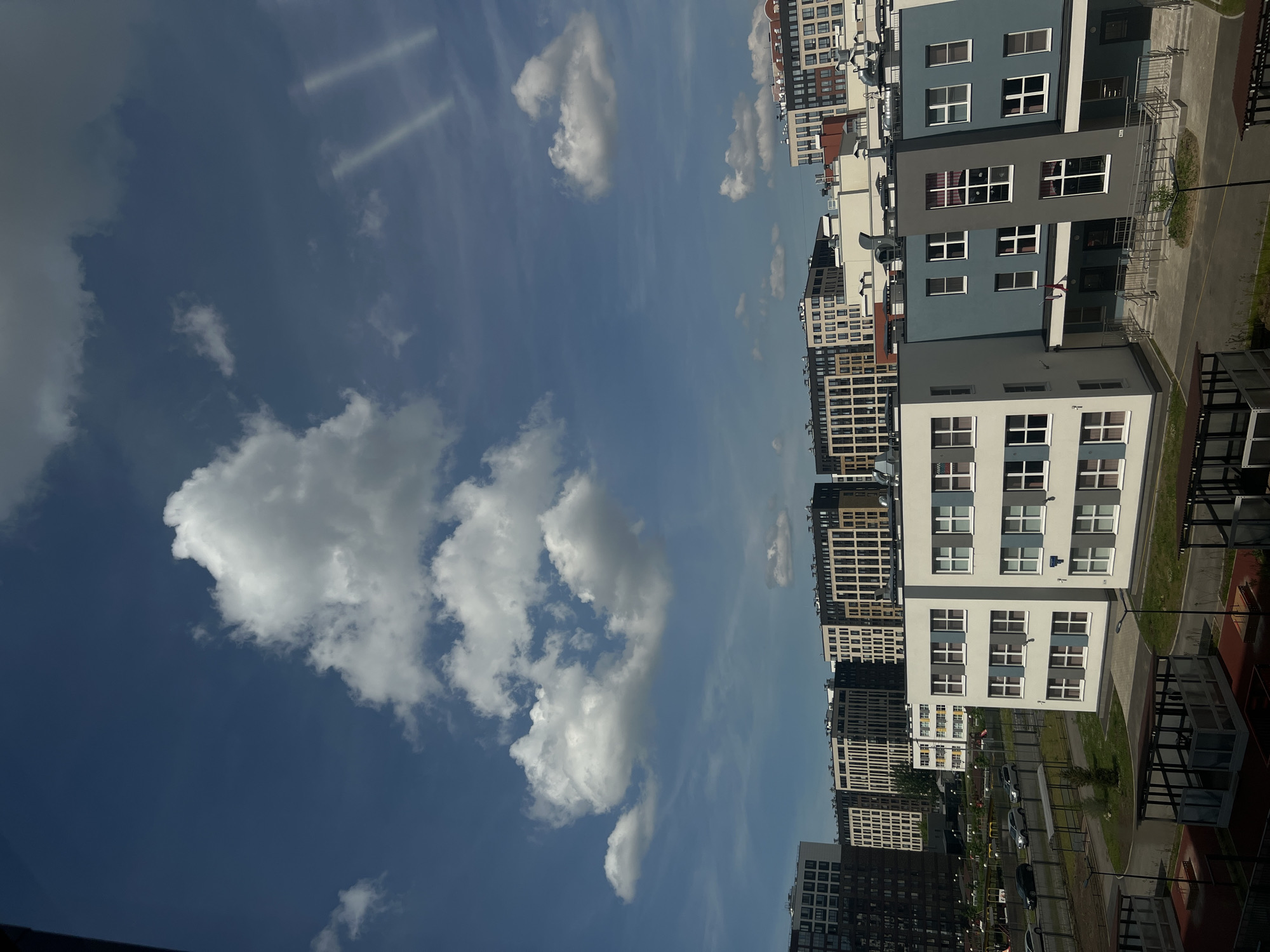The image shows a modern residential complex under a partly cloudy blue sky. In the foreground, there are children's play areas with covered structures. Behind these, a low-rise building with white and blue facades features multiple rows of windows. Further in the background, a series of taller residential buildings with a uniform, contemporary design, featuring many windows and balconies, dominate the skyline. Cars are visible on a road running alongside the complex, and some landscaping elements like trees and grass are present. The overall scene depicts a recently developed urban area.
