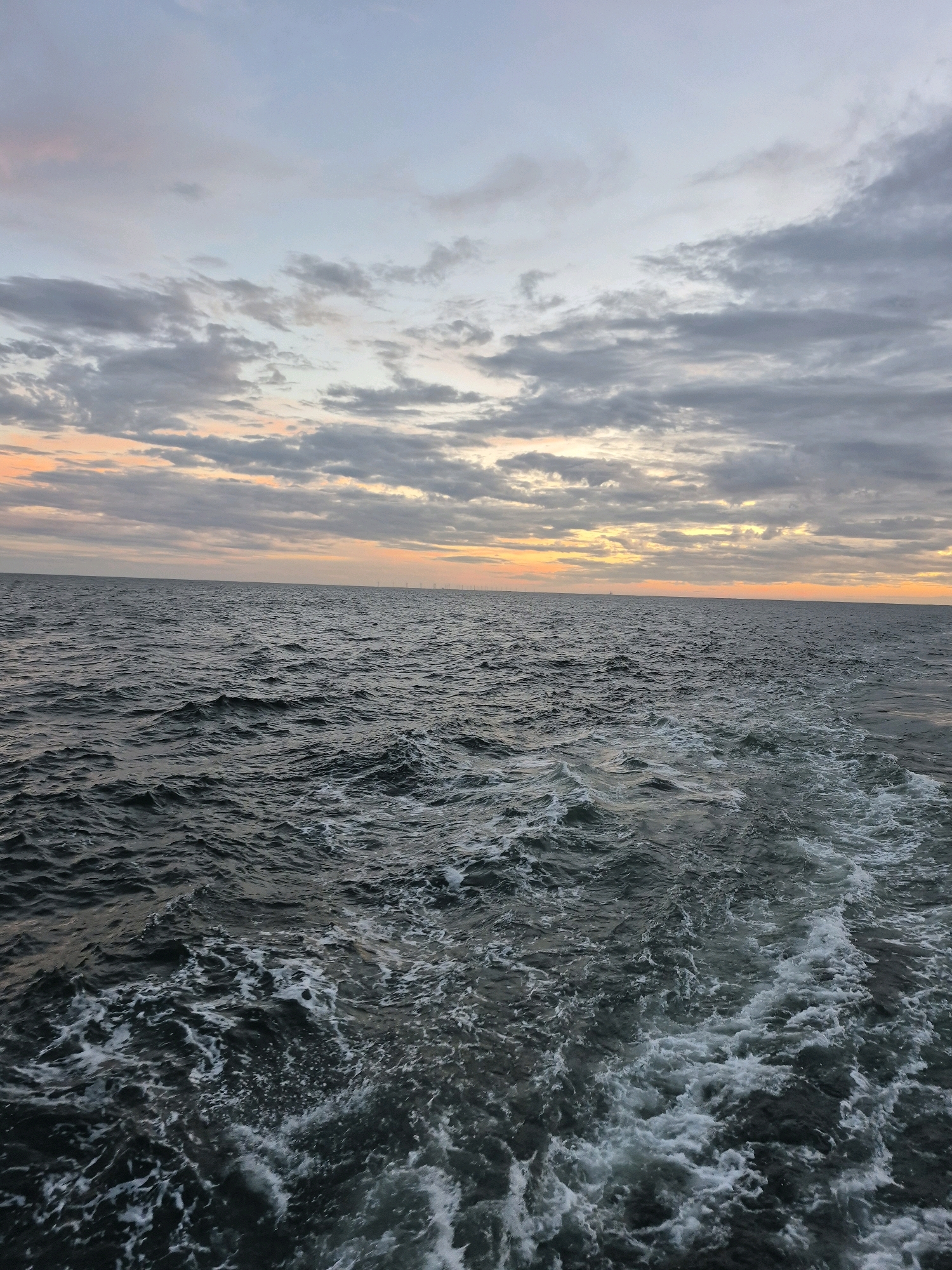 The image depicts a wide, dark expanse of choppy ocean water under a cloudy sky at dusk. The water is disturbed, with white foam and churned waves trailing towards the foreground, suggesting the wake of a boat. The sky is a mix of soft blues, grays, and subtle oranges and yellows near the horizon, indicating the last light of the day. In the far distance, a faint line of what appear to be wind turbines is visible on the horizon. The scene is devoid of people and human activity, focusing solely on the natural environment of the sea and sky. The location is described as Unknown, Germany.