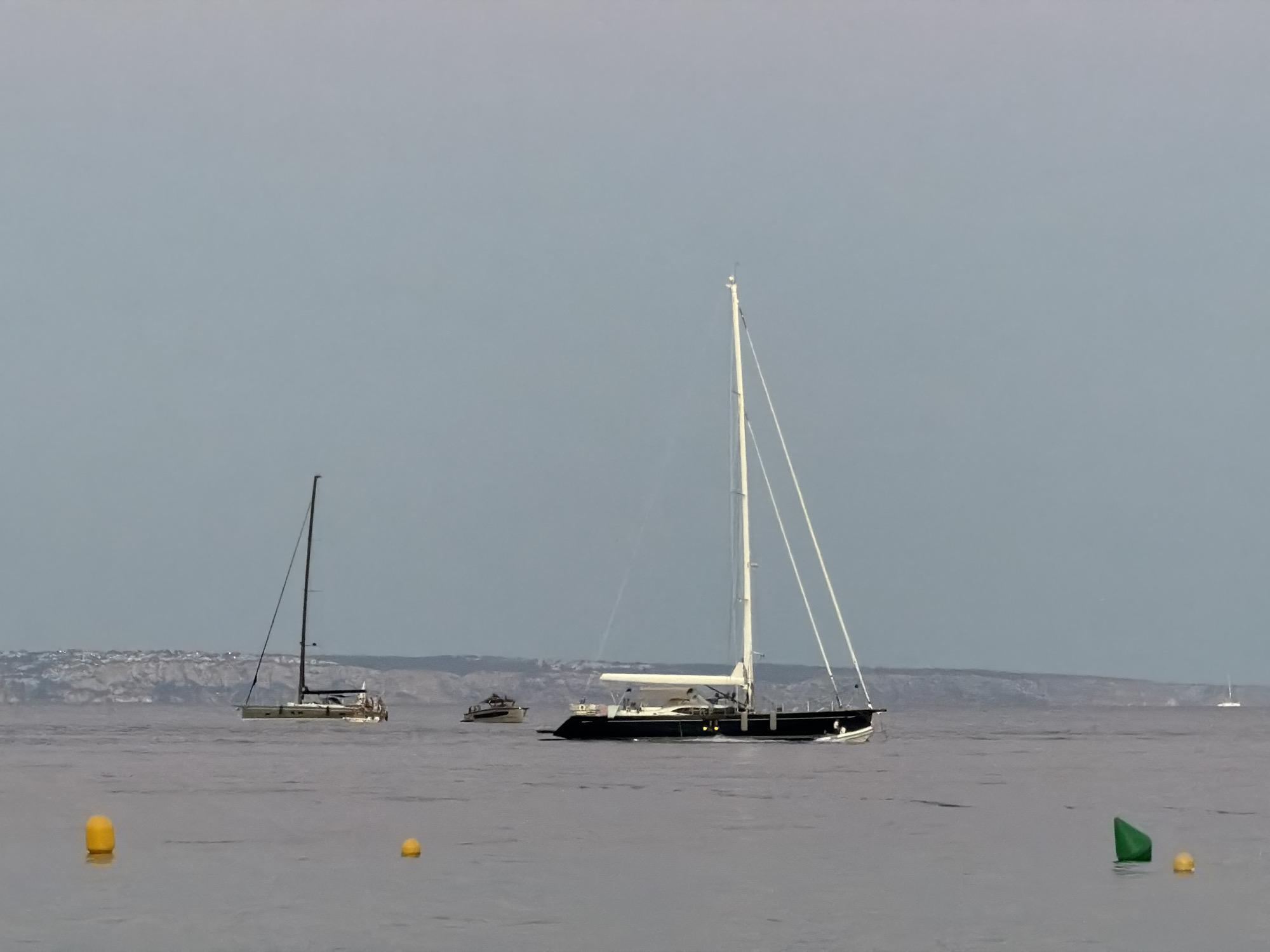 The image depicts a serene seascape with several sailboats and a distant coastline. In the foreground, two yellow buoys float on the calm water, with a green buoy visible to the right. Further out, a single-masted sailboat with a dark hull and white sails is prominently featured, appearing to be underway. To its left, another sailboat with a lighter hull and a single mast is also visible. In the middle ground, a small vessel with a dark hull and white superstructure is moving away from the viewer. The background reveals a hazy coastline with vegetation and structures, suggesting a Mediterranean setting. The sky is overcast with a pale blue hue. The scene appears to be set in or near the waters of Calvià, Spain, given the characteristic coastline and the type of vessels typically found in the Balearic Islands.