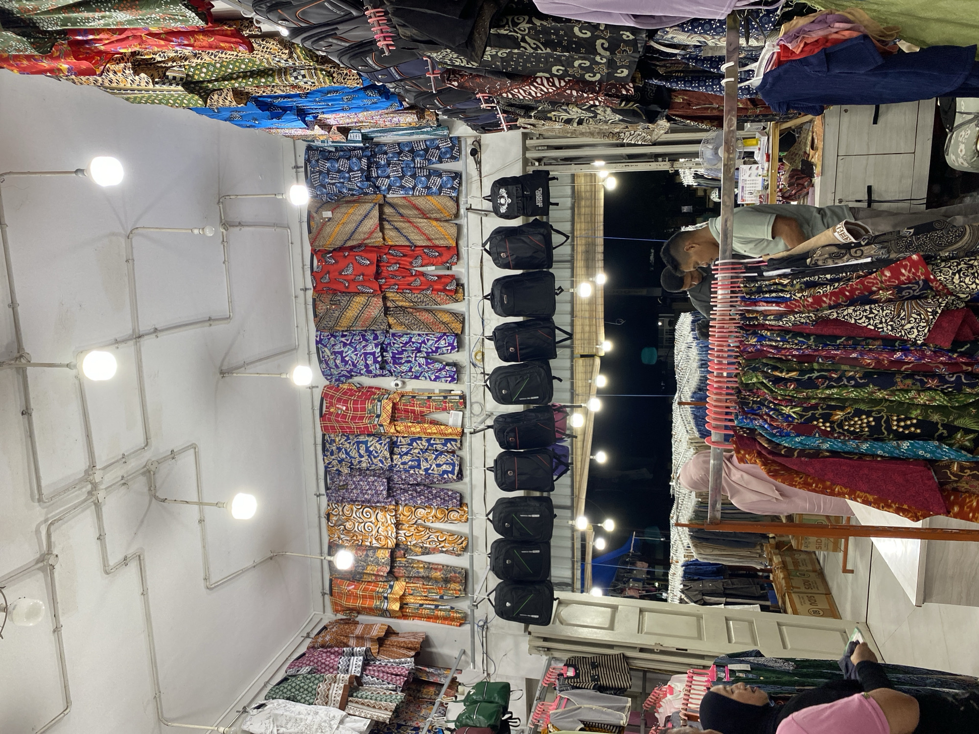 This media file shows an indoor market stall filled with various textiles and bags. The subjects include at least three people. One person, a man wearing a light blue polo shirt and dark pants, is examining textiles. Another person, a woman wearing a black hijab and a pink top, is looking at her phone while sitting. A third person, also wearing a black hijab, is partially visible in the foreground. The stall is brightly lit by overhead lights, and the textiles are displayed on racks, featuring colorful patterns, likely batik, common in Indonesia. Bags are also stacked on shelves and hung on walls.
The scene appears to be a retail environment, possibly a market or a shop. The setup suggests a commercial activity, with the people potentially being a vendor and customers. The overall atmosphere is that of a busy marketplace, though the exact time of day is not clearly discernible. However, the dark background visible through an opening suggests it might be evening or night. There are no overt weather cues.
Notable details include the abundance of intricately patterned fabrics, the various types of bags, and the electrical conduits running along the white ceiling. Some boxes with visible text, potentially shipping boxes, are stacked in the background. Although the text on the boxes is not entirely legible, one might contain the word "SOU". The interaction of the people with the goods implies shopping or selling activities are underway. The image conveys a sense of commerce and local culture, given the prominent display of batik-style fabrics. The location context of Lamteumen Barat, Indonesia, further reinforces the likely presence of such textile markets.