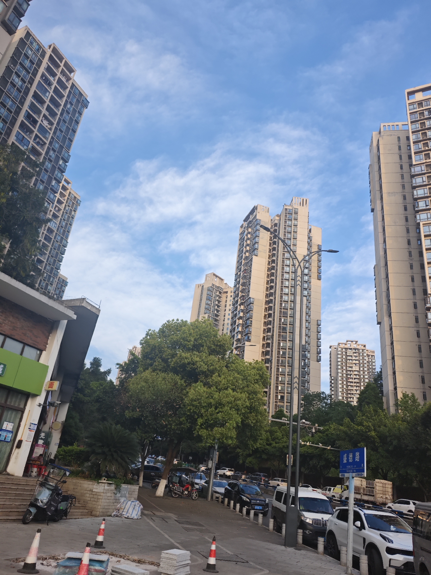 The image depicts a street scene in the Chongqing High-tech Zone, China. High-rise apartment buildings dominate the background, set against a blue sky with scattered clouds. In the foreground, a street with parked cars lines the right side. A row of trees separates the street from the buildings in the background. On the left, there is a storefront with a green sign and a set of stairs leading up to it. A scooter is parked near the stairs. There are road work cones and stacked tiles on the side of the street. A blue street sign with Chinese and English writing is visible amidst the trees and parked cars, seemingly indicating the name of the road.