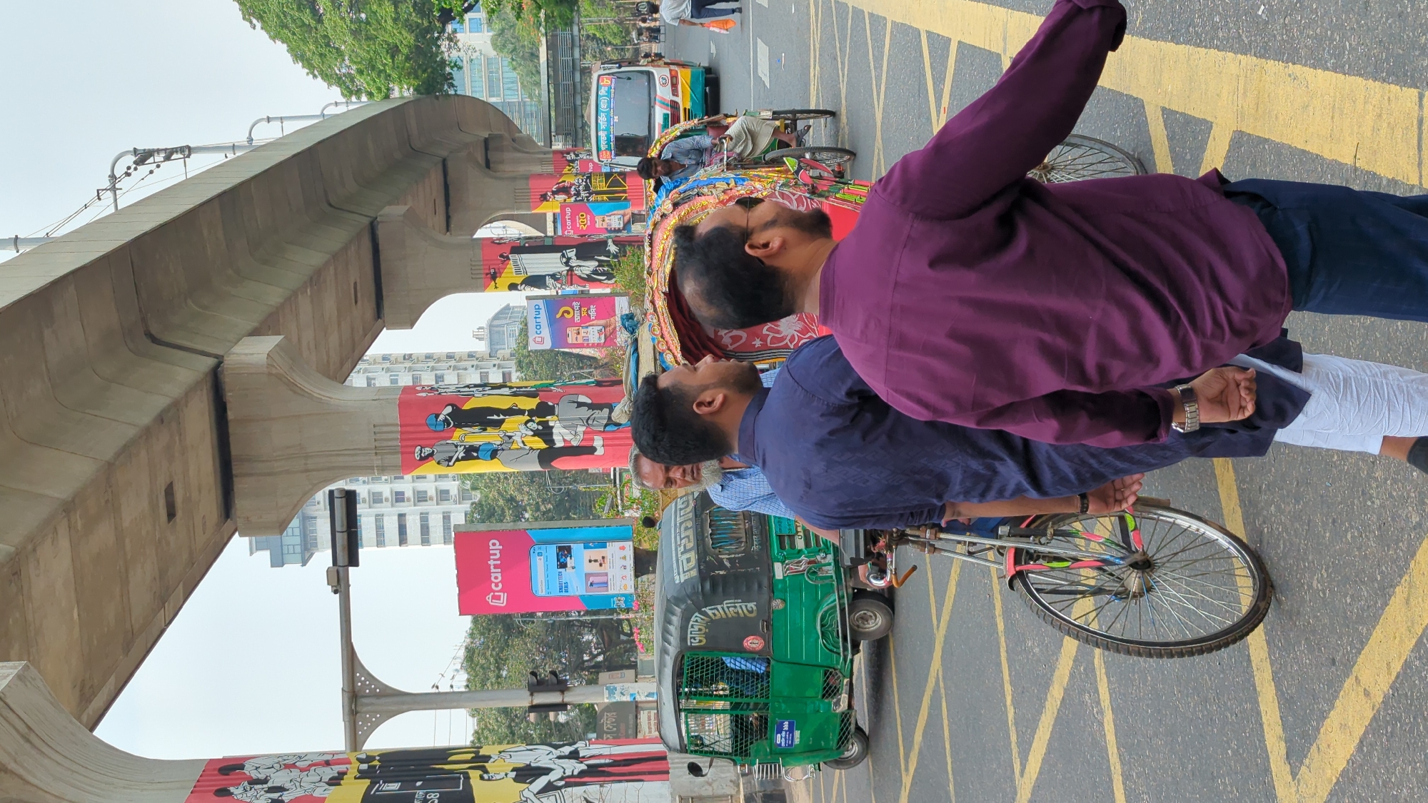 The image depicts a bustling urban street scene in Dhaka, Bangladesh, under what appears to be a clear daytime sky. The perspective is from the street level, looking towards an elevated concrete structure.
In the foreground, two men stand on the asphalt road, their backs slightly turned, both looking upwards. The man on the right wears a deep purple shirt and dark trousers, while the man on the left is in a navy blue shirt and white pants, with a metallic wristwatch visible on his left wrist. The road beneath them has prominent yellow crosshatch markings.
To the left of the foreground subjects, a colorful cycle rickshaw with its puller is visible, positioned near a green auto-rickshaw. Further down the street, various vehicles including another auto-rickshaw and a bus with Bengali text on its side are present, alongside other pedestrians.
Throughout the scene, tall poles adorned with advertising banners line the street. These banners frequently feature the "cartup" logo, images of electronic devices like smartphones, and Bengali text. One large banner also displays stylized monochrome figures.
Dominating the mid-ground and background is a massive elevated concrete structure, likely part of a metro line or flyover, supported by robust pillars. This structure runs parallel to the street. In the distance, modern high-rise buildings and some trees are visible against the bright sky. Bengali text is also discernible on the green auto-rickshaw ("চালিত" - chalito, meaning 'driven' or 'operated') and on the distant bus, indicating routes or company names. The overall atmosphere suggests a vibrant and active city environment with a mix of traditional transport and modern infrastructure.