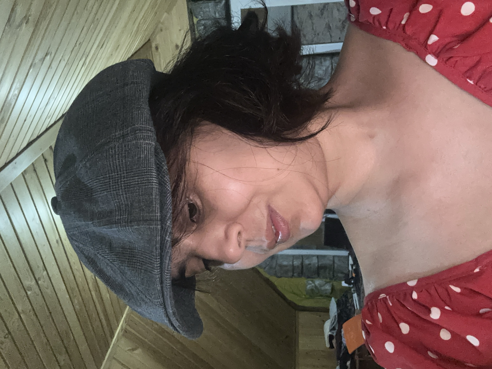 A medium shot captures a woman wearing a plaid flat cap and a red polka dot dress. She is looking down and to her left, with her mouth slightly open. Her dark hair is cut short and appears to be slightly tousled. The background features a wooden ceiling and a stone wall. The scene is indoors, possibly in an attic or loft space. There are no discernible elements in the image that would indicate the location within Kalininskaya, Russia.