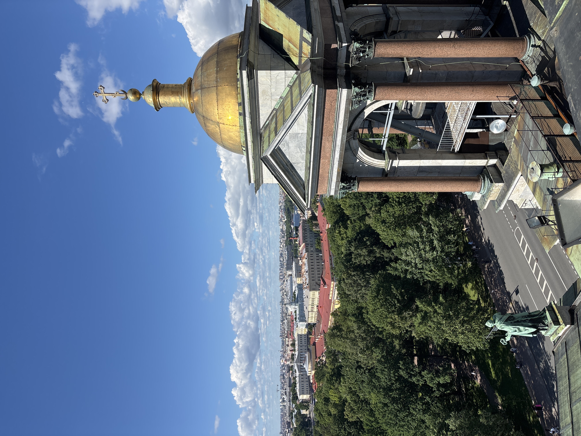 This is an aerial view from the colonnade of Saint Isaac's Cathedral in Saint Petersburg, Russia, looking northeast. In the foreground, the gilded dome of the cathedral is visible, topped with a cross. The dome sits atop a structure with large, reddish-brown columns and white marble facing. Below this, a large green statue of a figure holding a wreath stands on a pedestal near a road.
Beyond the cathedral's structure, a dense canopy of green trees stretches across the midground, with several buildings with red roofs peeking through. Further into the distance, the cityscape of Saint Petersburg unfolds, featuring numerous buildings with light-colored facades and darker roofs. The horizon is dominated by a clear blue sky with scattered white cumulus clouds. Sunlight illuminates the scene, creating shadows and highlighting the golden dome. Several people are visible on the pathways and sidewalks below.