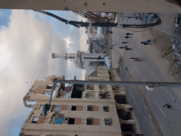 The image depicts an elevated view of a street in Khan Yunis, Palestinian Territory, showing an urban environment marked by extensive damage. On the left, a multi-story building with a light-colored facade exhibits severe structural damage, including a collapsed upper facade, exposed rebar, and numerous empty or broken windows, with some openings covered by blue tarpaulins.
In the middle ground, a white mosque featuring a prominent dome and a tall, cylindrical minaret stands, also displaying visible damage to its exterior. Beyond the mosque, additional buildings appear heavily damaged or reduced to rubble.
The paved street below has visible lane markings and is populated by approximately fifteen individuals walking in various directions. Most are adults, and some carry bags or bundles. One individual is observed squatting on the road in the immediate foreground, near a street light pole. Rubble and twisted rebar are scattered along the right edge of the road in the foreground. Utility cables are visible attached to the exterior of a building on the far right of the frame, from which the photograph appears to have been taken. The sky above is partly cloudy.