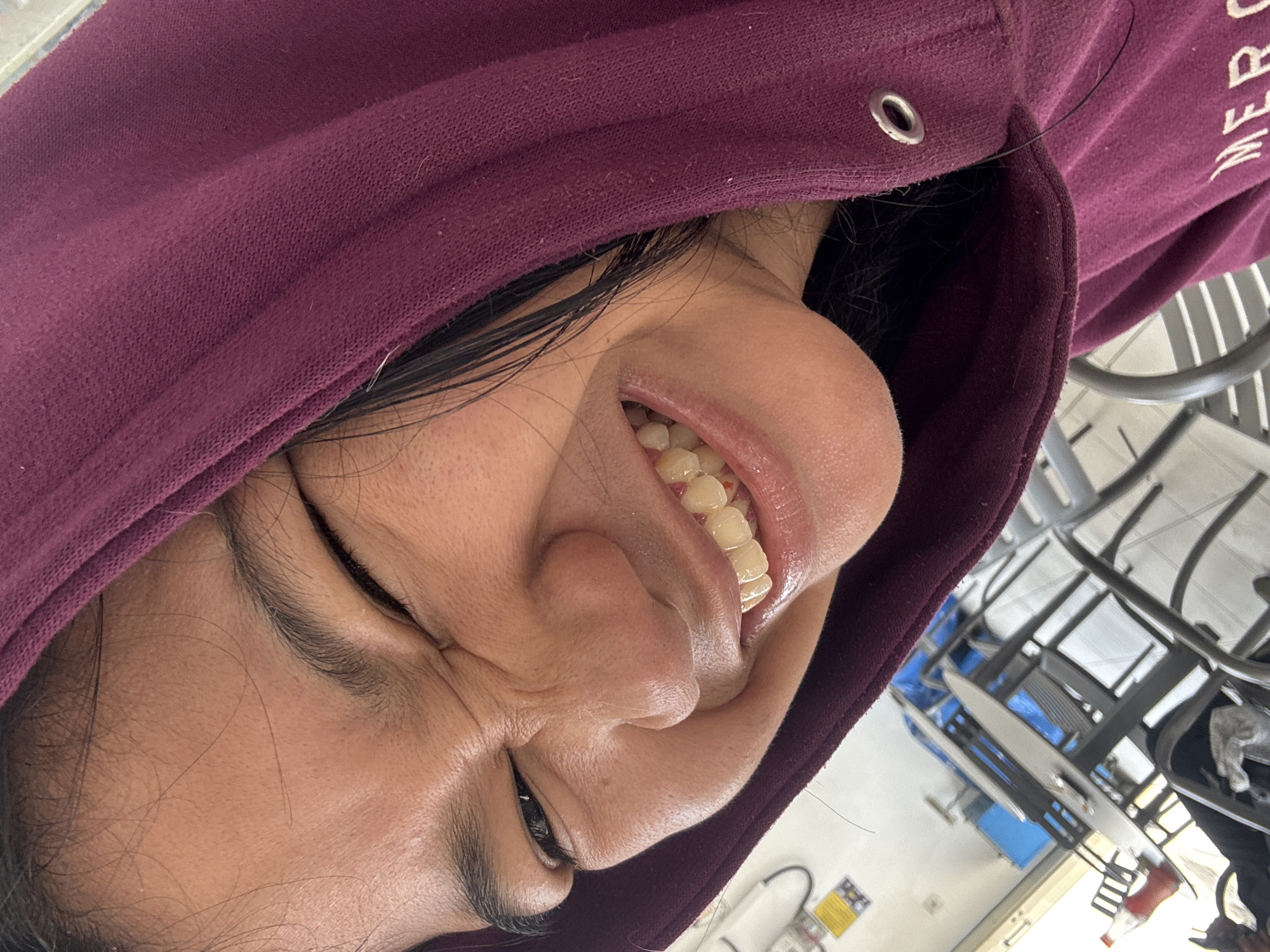 A close-up, low-angle, self-portrait shows a person with dark hair, wearing a maroon hoodie, and making a grimacing facial expression with their eyes squinted shut and mouth open, revealing their teeth. The person is positioned in what appears to be an outdoor seating area with tables and chairs, possibly in a food court or cafe. The background shows tables with chairs, some covered with blue tarps, and a few bottles on a table. The overall setting suggests a casual, possibly commercial, environment. The lighting indicates daytime. The image does not contain any information about the specific location in North Jakarta, Indonesia.