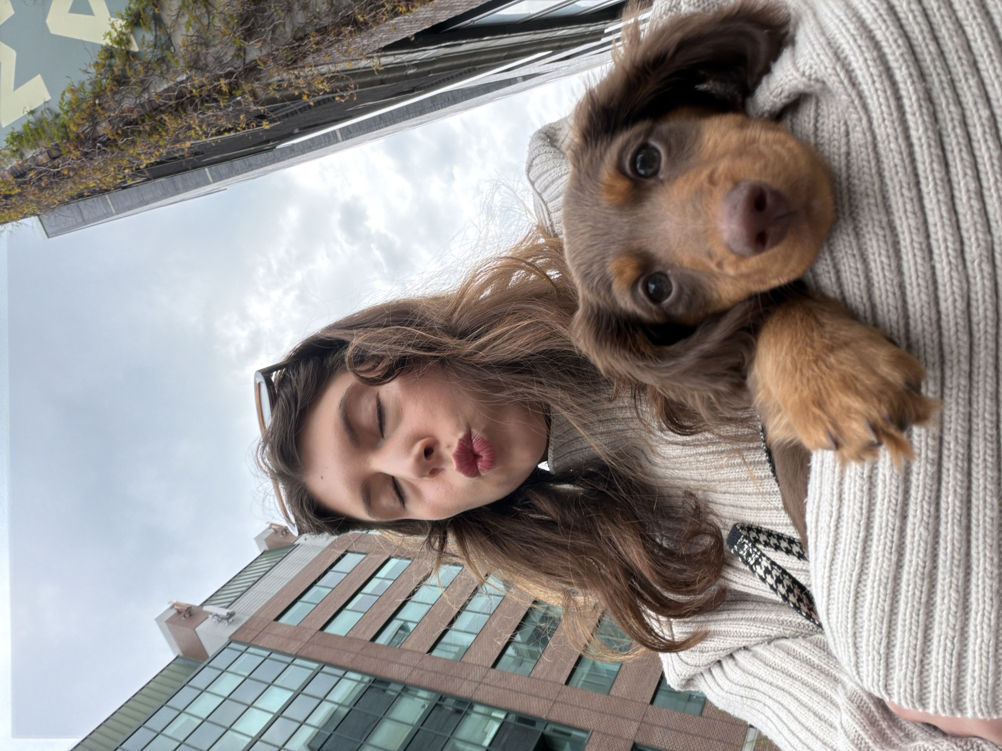 A young woman with long brown hair is holding a small, brown and tan dachshund puppy. The woman has her eyes closed and her lips pursed as if to give a kiss. She is wearing a light-colored sweater and sunglasses are perched on her head. The dog is nestled against her chest with its head visible, looking at the camera. In the background, there is a building with a glass facade, partially visible, and a section of a building with a green, vine-covered wall. The photograph appears to have been taken from a low angle looking upward. It is not possible to verify if the location is in the city of Montreal, Canada based on the current image.