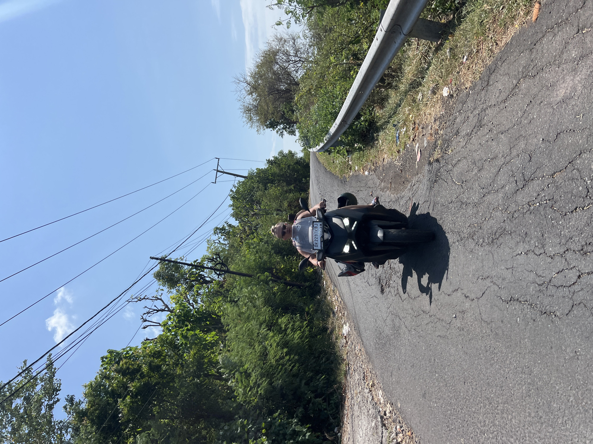 In the city of Abang, Indonesia, a person rides a black scooter along a cracked asphalt road. The rider, wearing a grey tank top and sunglasses, is positioned in the center of the frame, facing forward. The scooter has a clear windshield, and a license plate displaying "DK2291KBJ". The road curves to the right and is bordered by a metal guardrail on the right side. Lush green trees and vegetation line both sides of the road, with a bright blue sky overhead, punctuated by telephone wires. The scene appears to be during the daytime with ample sunlight.