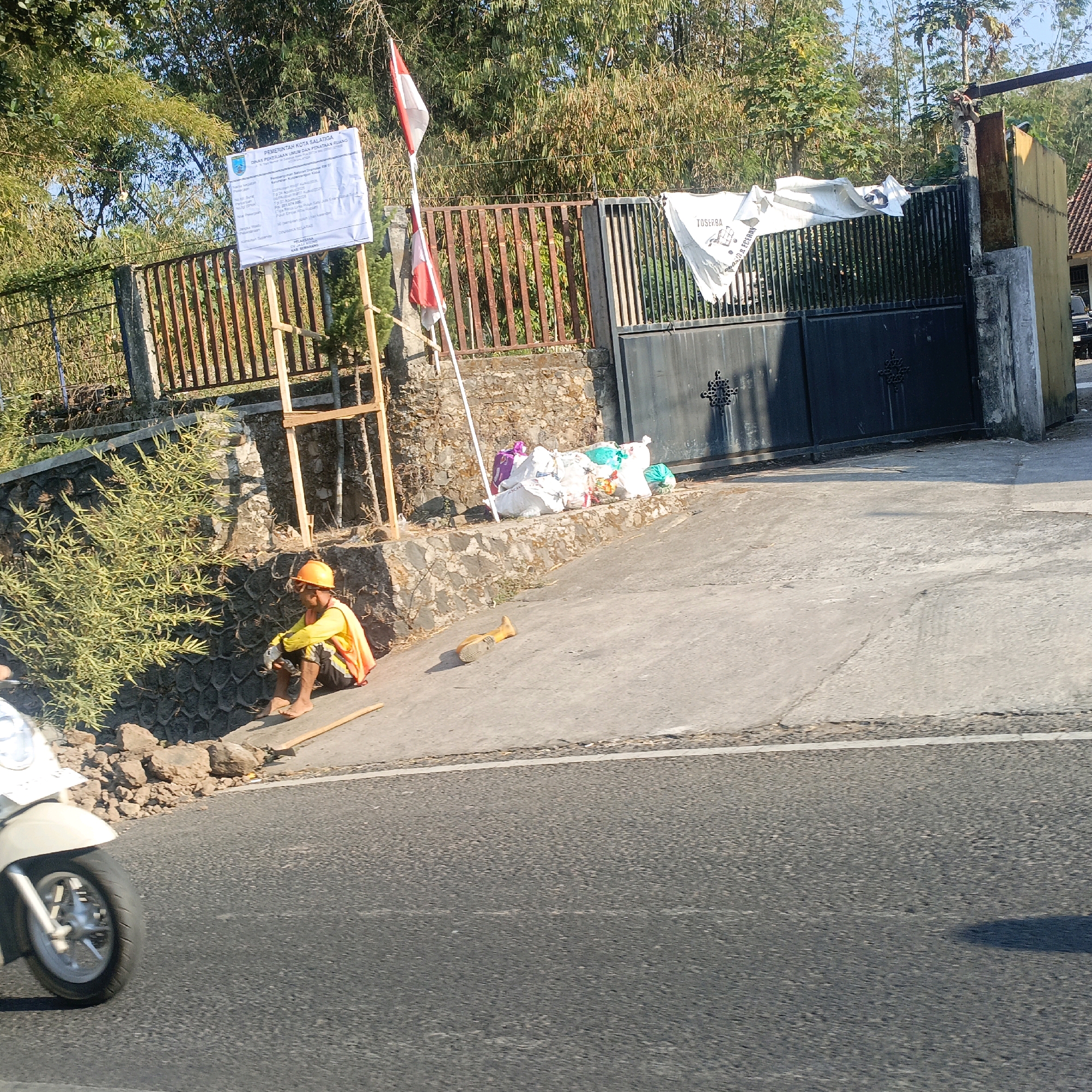 The image depicts a scene on a roadside in Salatiga, Indonesia. A construction worker wearing a hard hat and a safety vest is seated on the curb next to a stone wall. Piles of white and colored sacks are stacked on the ground near a metal gate. A white scooter is partially visible in the foreground, moving on the asphalt road. In the background, a sign is displayed on a wooden frame, and Indonesian flags are also present. Trees and vegetation surround the area. The sun is casting shadows, indicating it is daytime.
